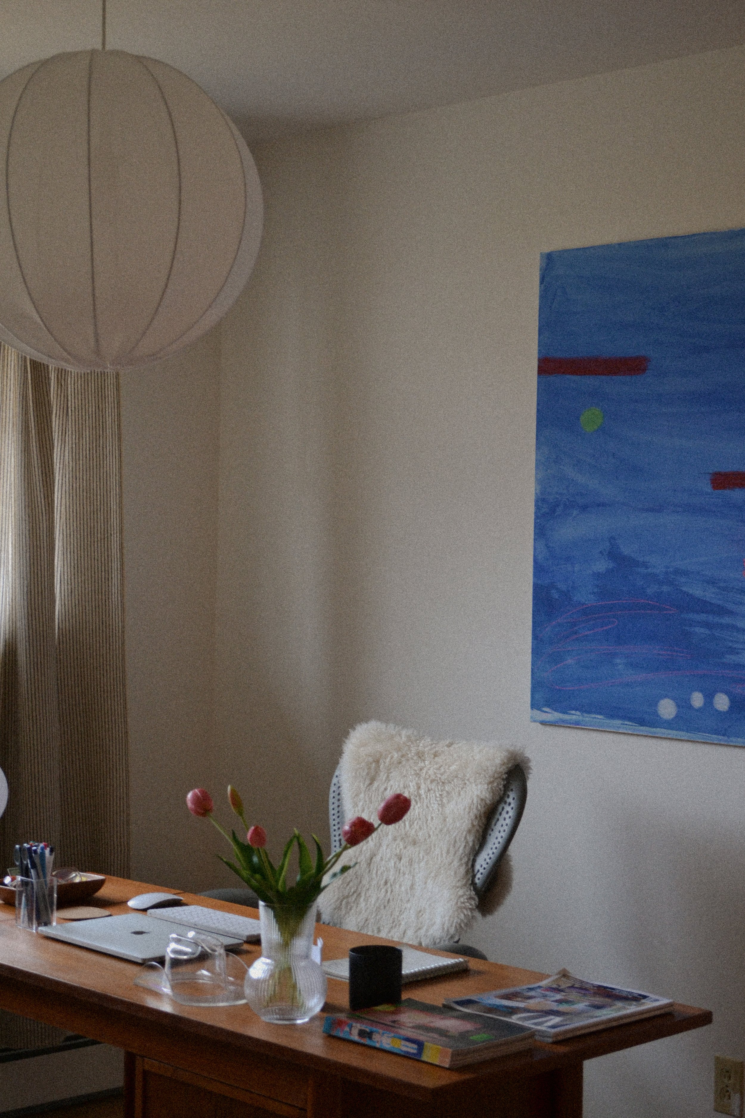 Home office with wooden desk, laptop, vase with tulips, notebooks, and abstract artwork on wall.