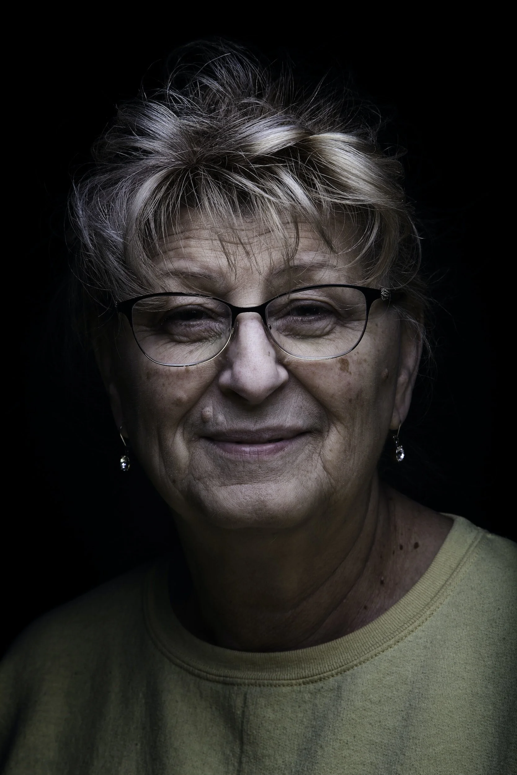 Close-up portrait of an elderly woman with short, curly blonde hair, wearing glasses, earrings, and a light-colored shirt, smiling against a black background.