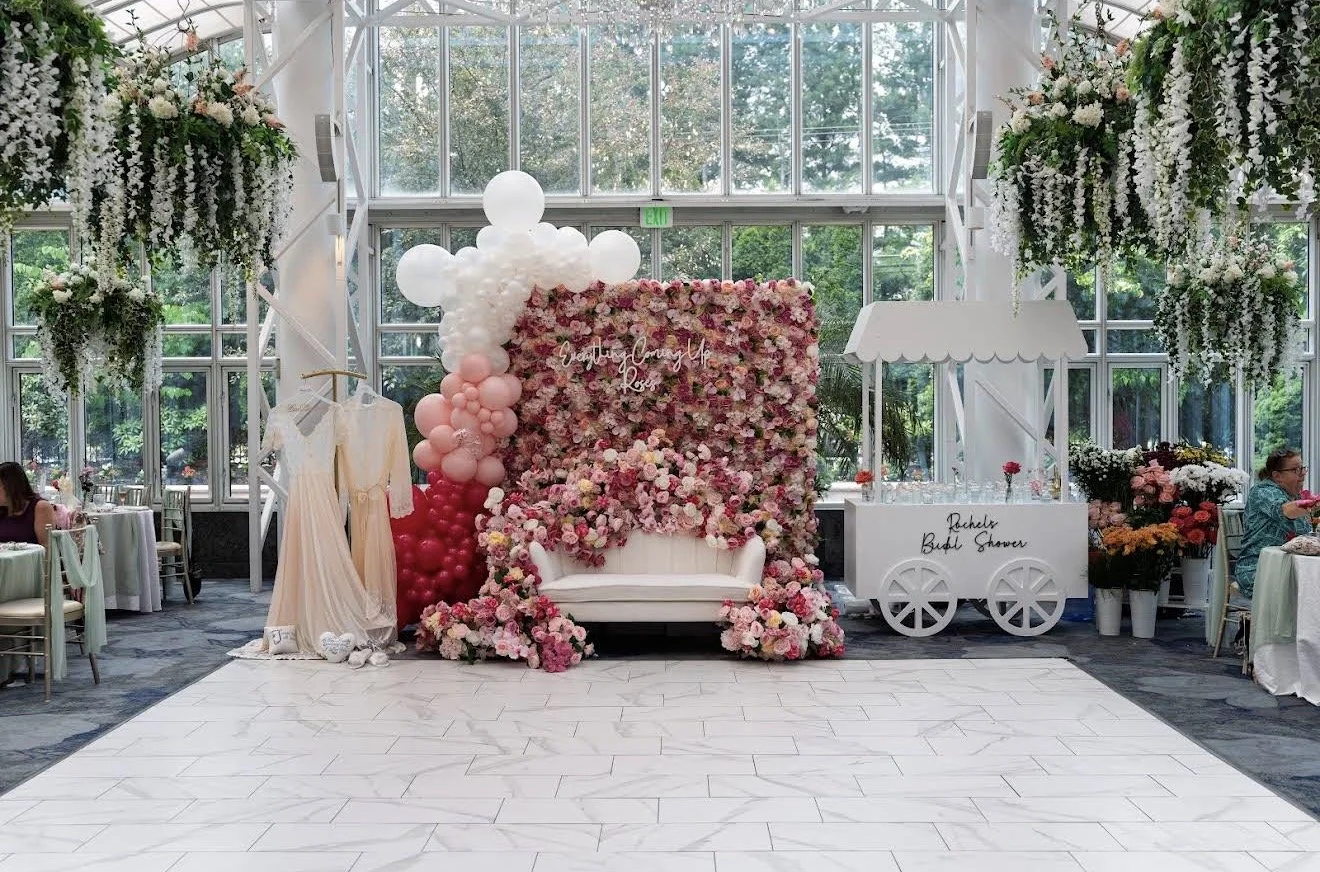 Indoor wedding or celebration setting with large floral backdrop, balloons, and lounge area, featuring tables and chairs, and decorated with flowers and greenery.