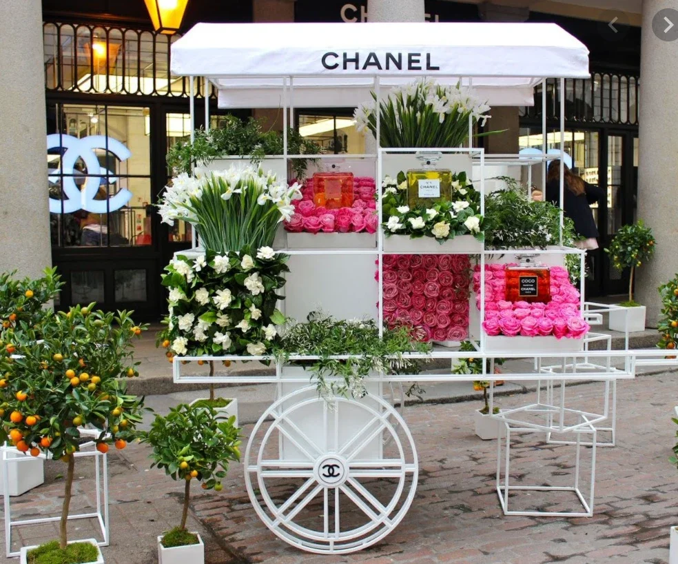 A white floral cart with a canopy labeled 'CHANEL' displays various arrangements of pink and white flowers, with greenery, in front of a storefront with large windows and a Chanel logo.