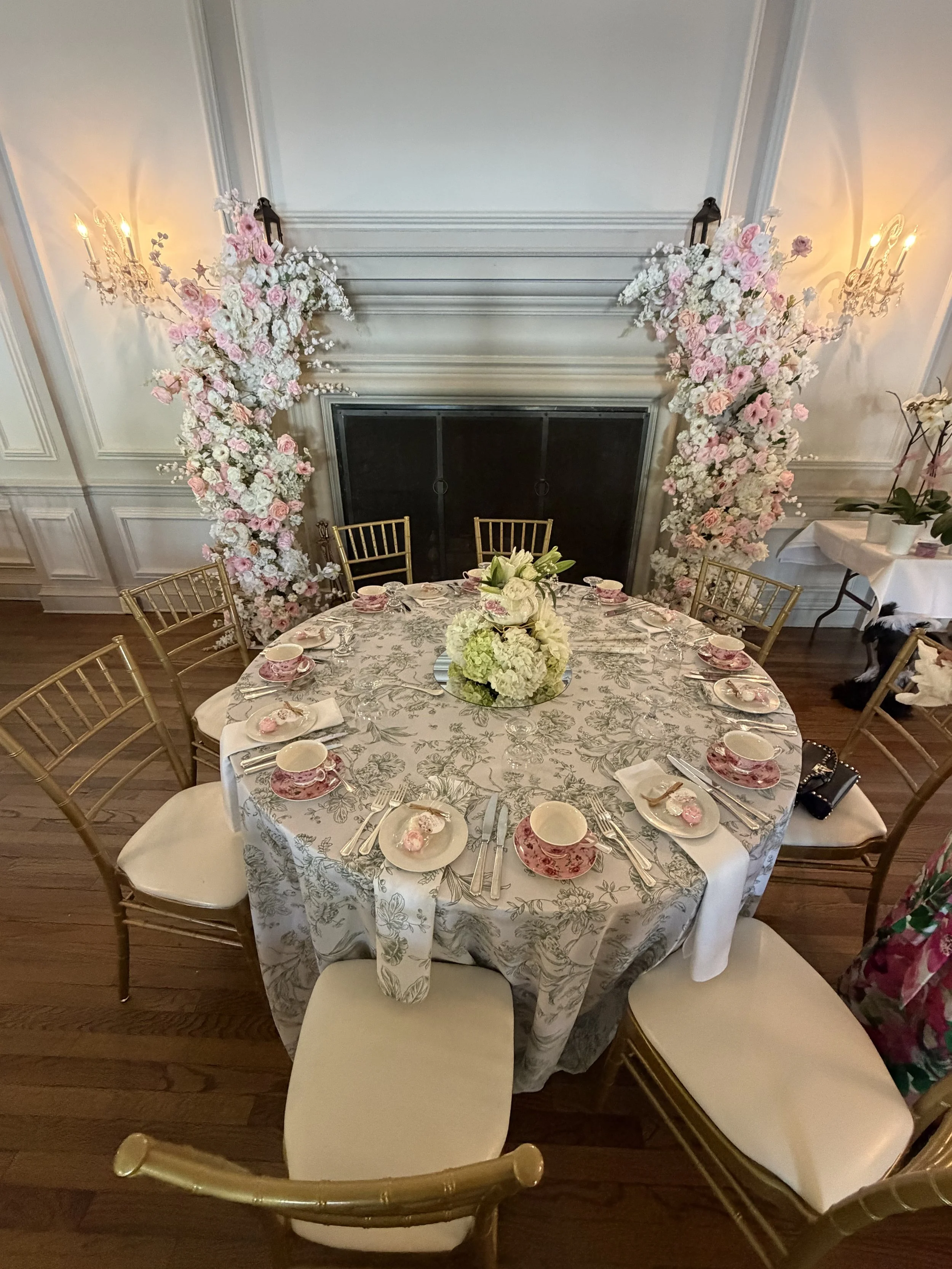 A round dining table with a floral patterned tablecloth, a floral centerpiece, and place settings including pink and white china, silverware, and glasses. The table is surrounded by gold and white chairs. Behind, there is a floral arch made of pink a