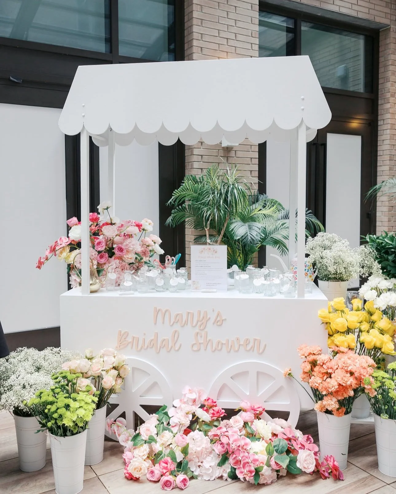 Decorative cart with a white canopy and pink sign reading 'Mary's Bridal Shower', surrounded by pink, white, yellow, and peach flowers in vases.