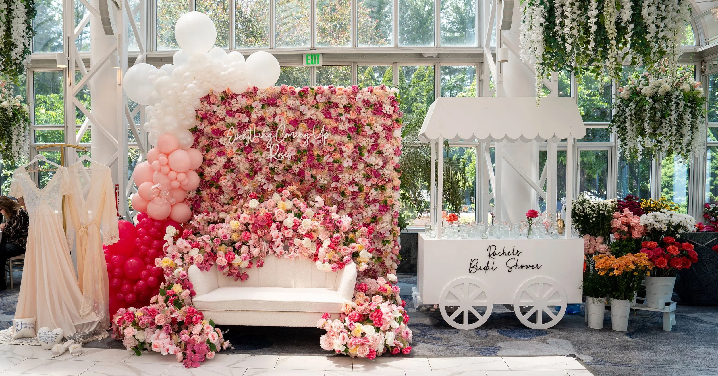 A floral backdrop decorated with pink, white, and cream roses, with a balloon arch and a white loveseat in front. To the right is a white cart with a sign that reads 'Rachels Bridal Shower' and various flowers. To the left are dresses and a woman sitting. The setting is a bright space with large windows and greenery outside.