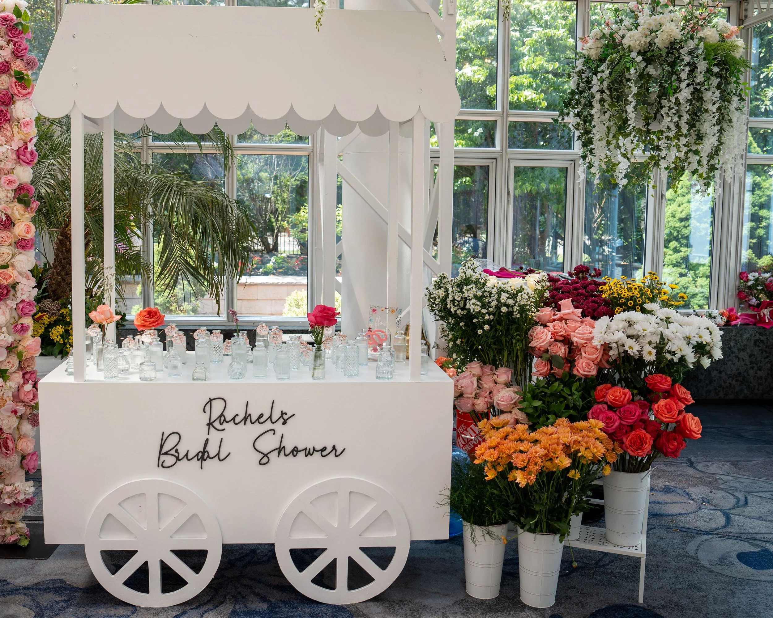 White flower cart decorated for a bridal shower with a sign that reads "Rachels Bridal Shower." The cart is surrounded by various colorful flowers, including roses and daisies, in vases and buckets. There are large windows in the background letting i