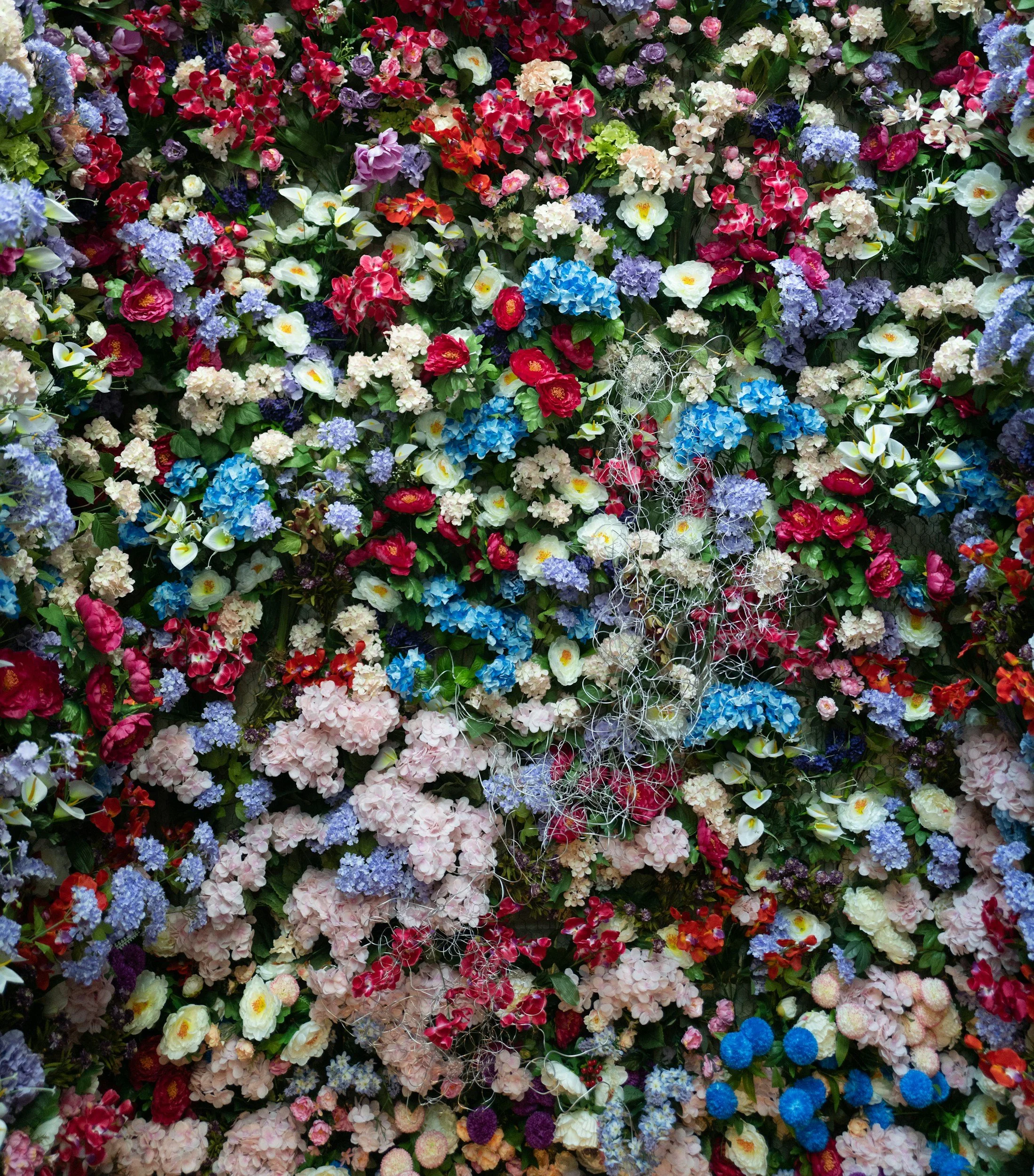 Colorful wall of various artificial flowers, including hydrangeas, roses, lilies, and other blooms in pink, purple, blue, white, and red. permanent installations in nj ny floral murals flower walls permanent b2b