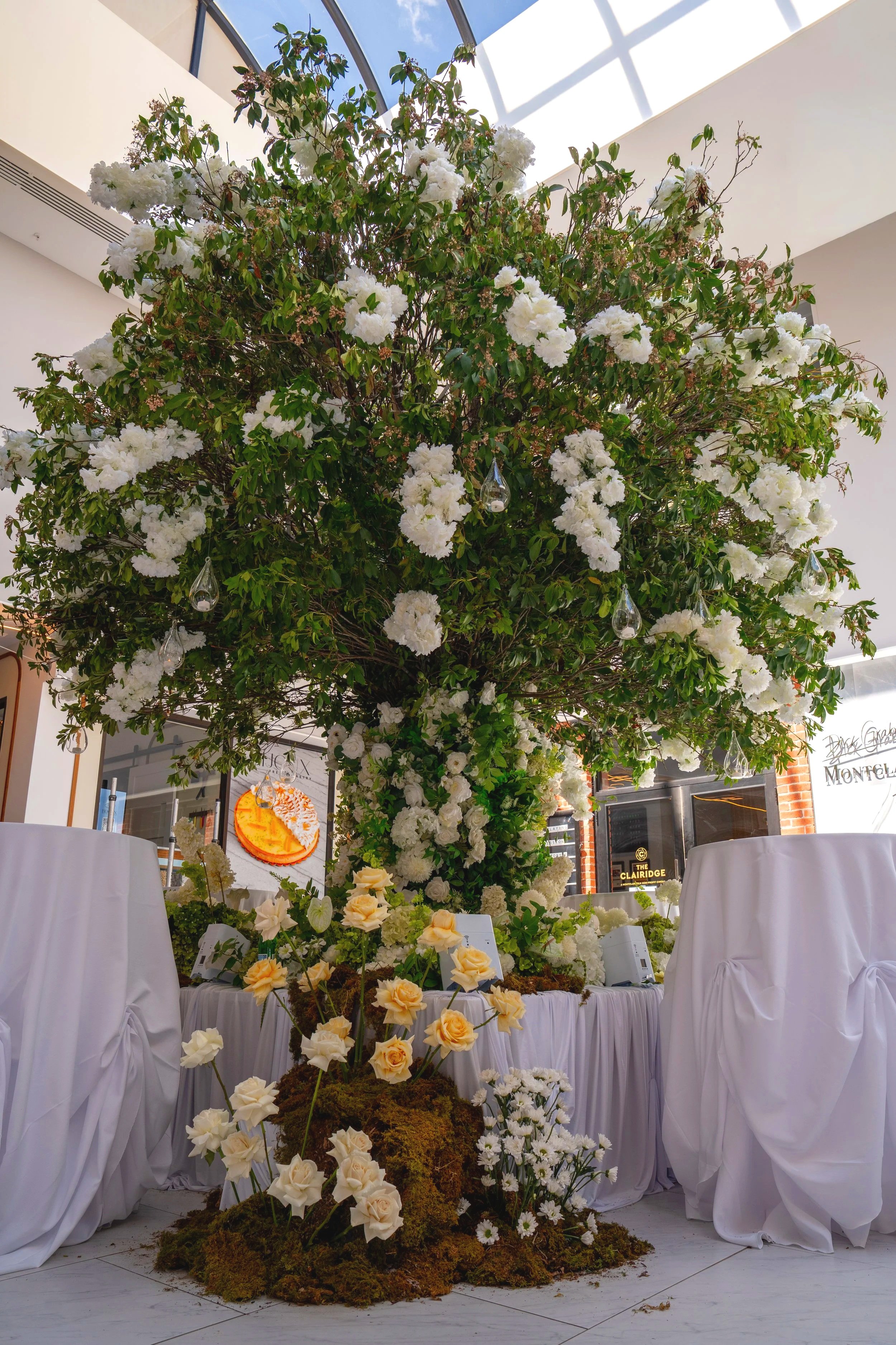 A large indoor floral display featuring a tall tree with white flowers, surrounded by smaller yellow and white flowers on moss and ground cover, with white draped tables and signs at a decorated event or venue.