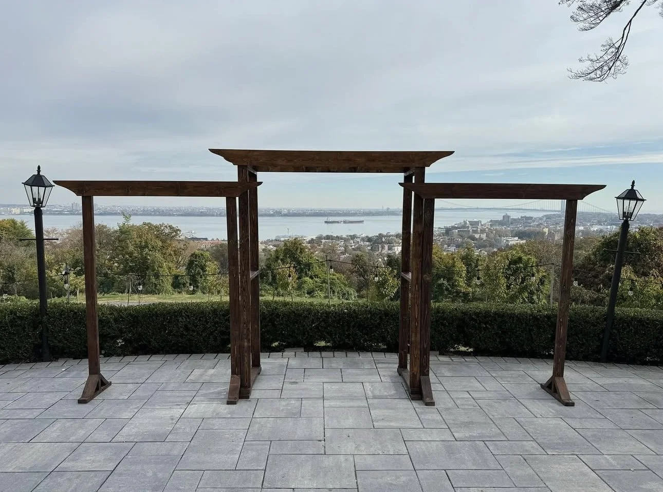 Wooden archway on a tiled patio with a city and river view in the background, flanked by street lamps on each side. NYC skyline ceremony