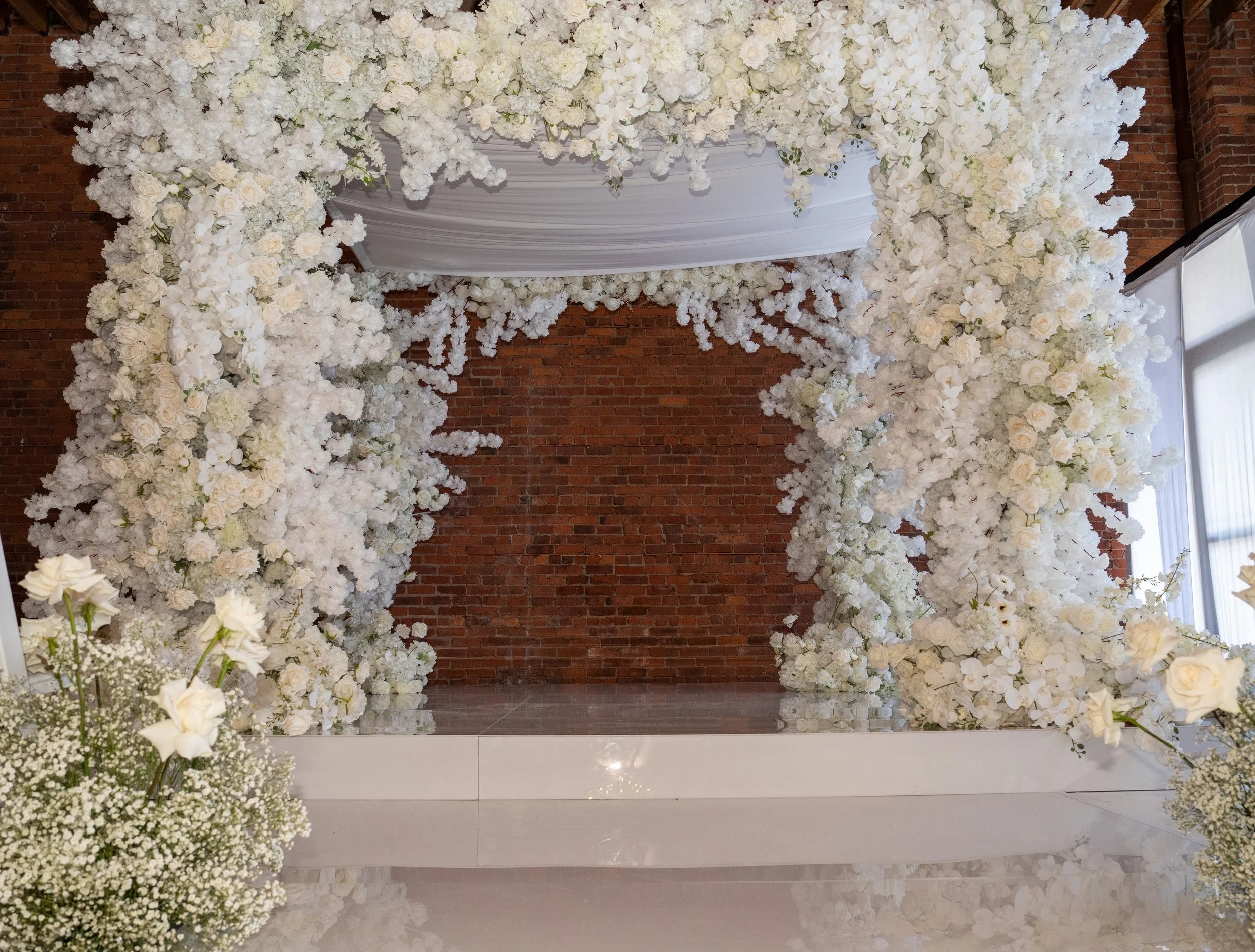 Elegant wedding altar decorated with white flowers and draped fabric, set against a brick wall, with natural light streaming in. Chuppah design in nj luxury weddings florals in montclair nj 