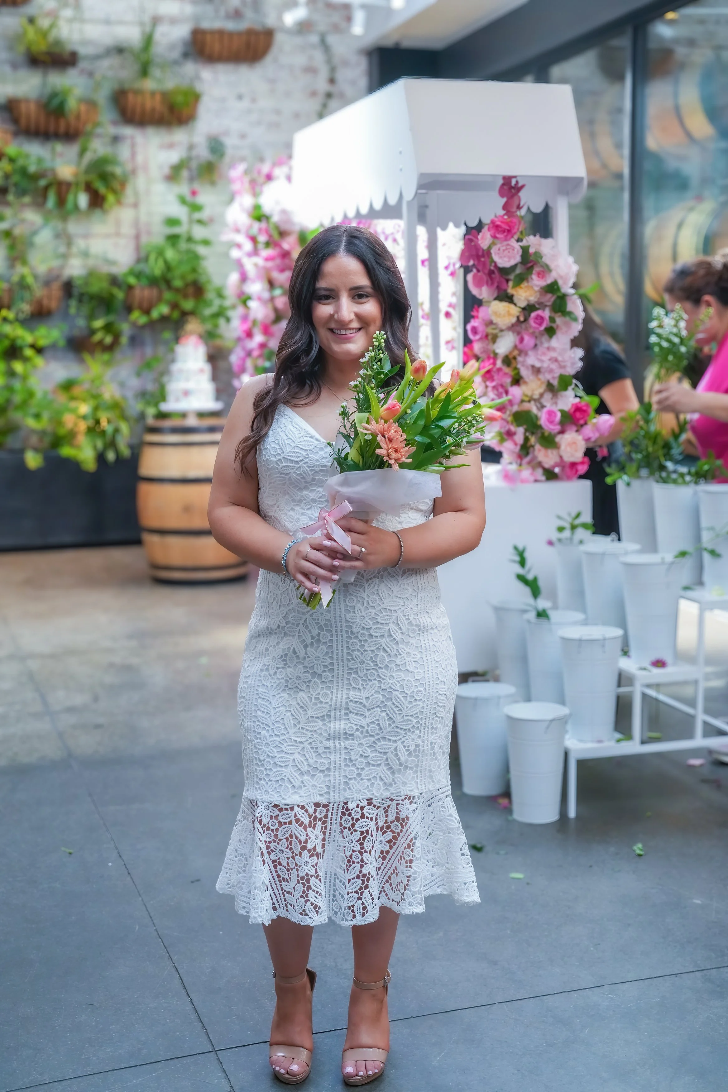 A woman in a white lace dress holding a bouquet of pink and peach flowers, standing in front of a floral arrangement and pink flower decorations at an event or celebration.