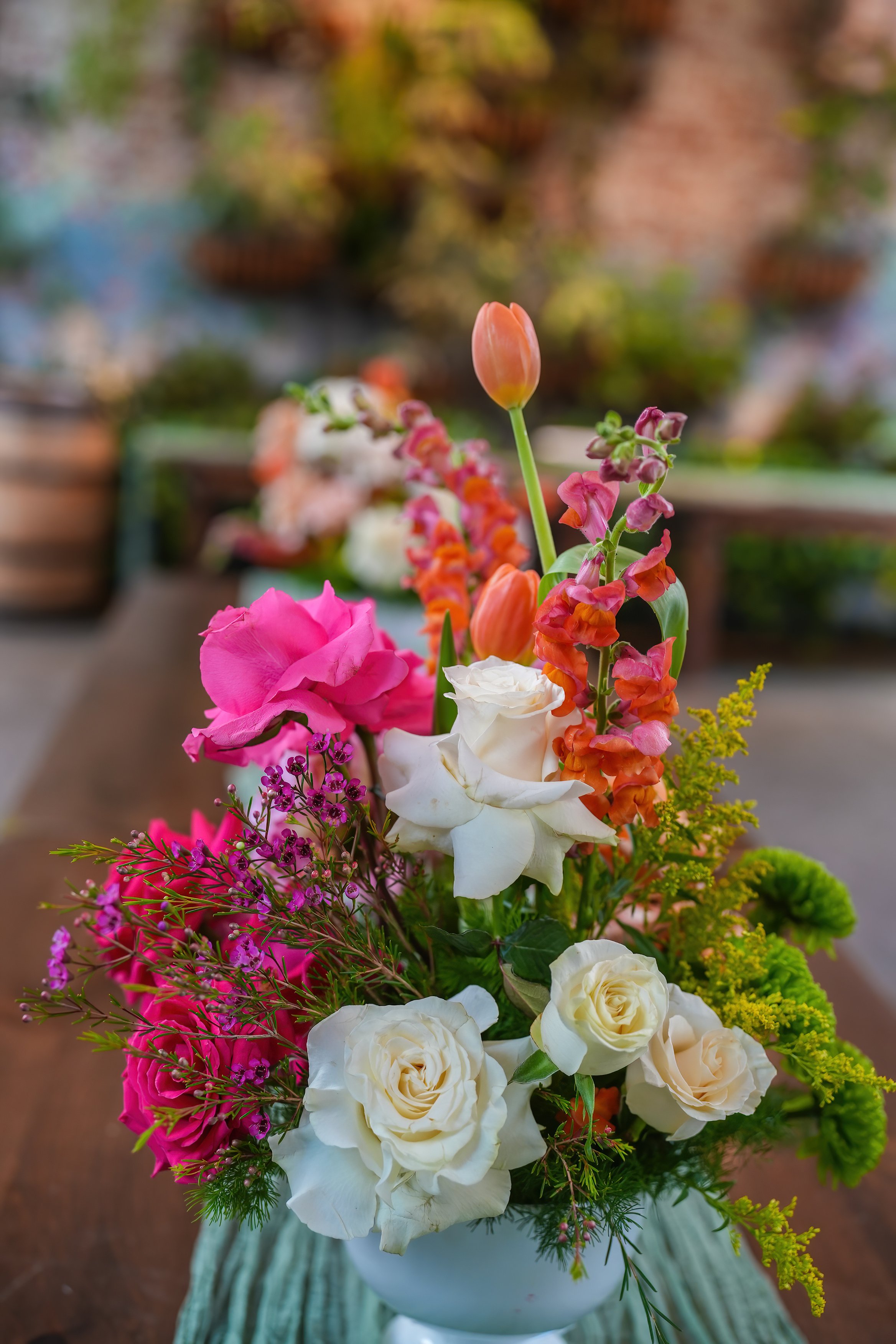 Colorful flower arrangement in a white vase, featuring pink, white, orange, and yellow flowers, placed on a striped cloth on a wooden surface.