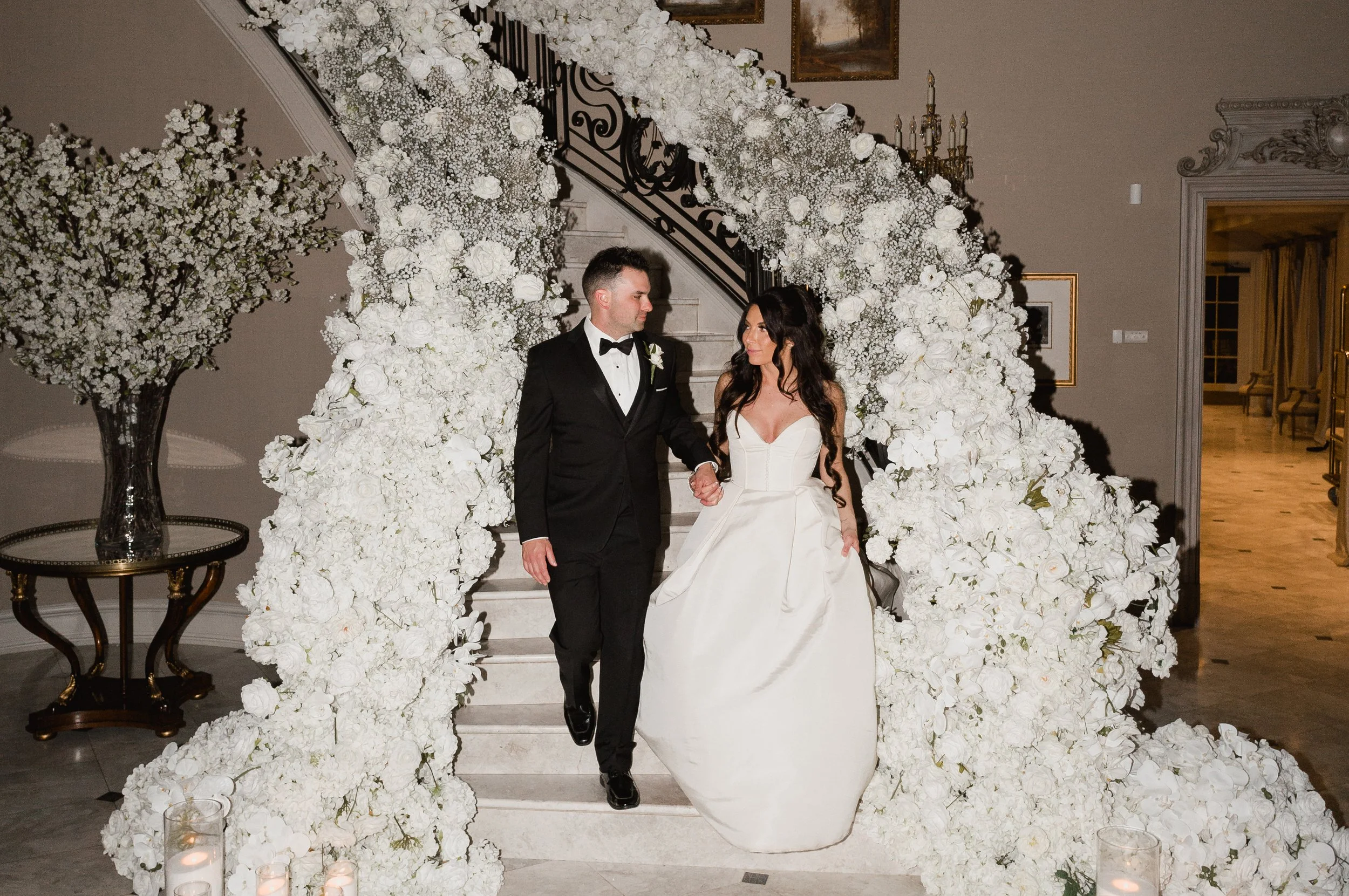 A bride and groom holding hands on a staircase surrounded by white floral arrangements at their wedding. Park chateau NJ wedding venue luxury stair case floral wrap with candles