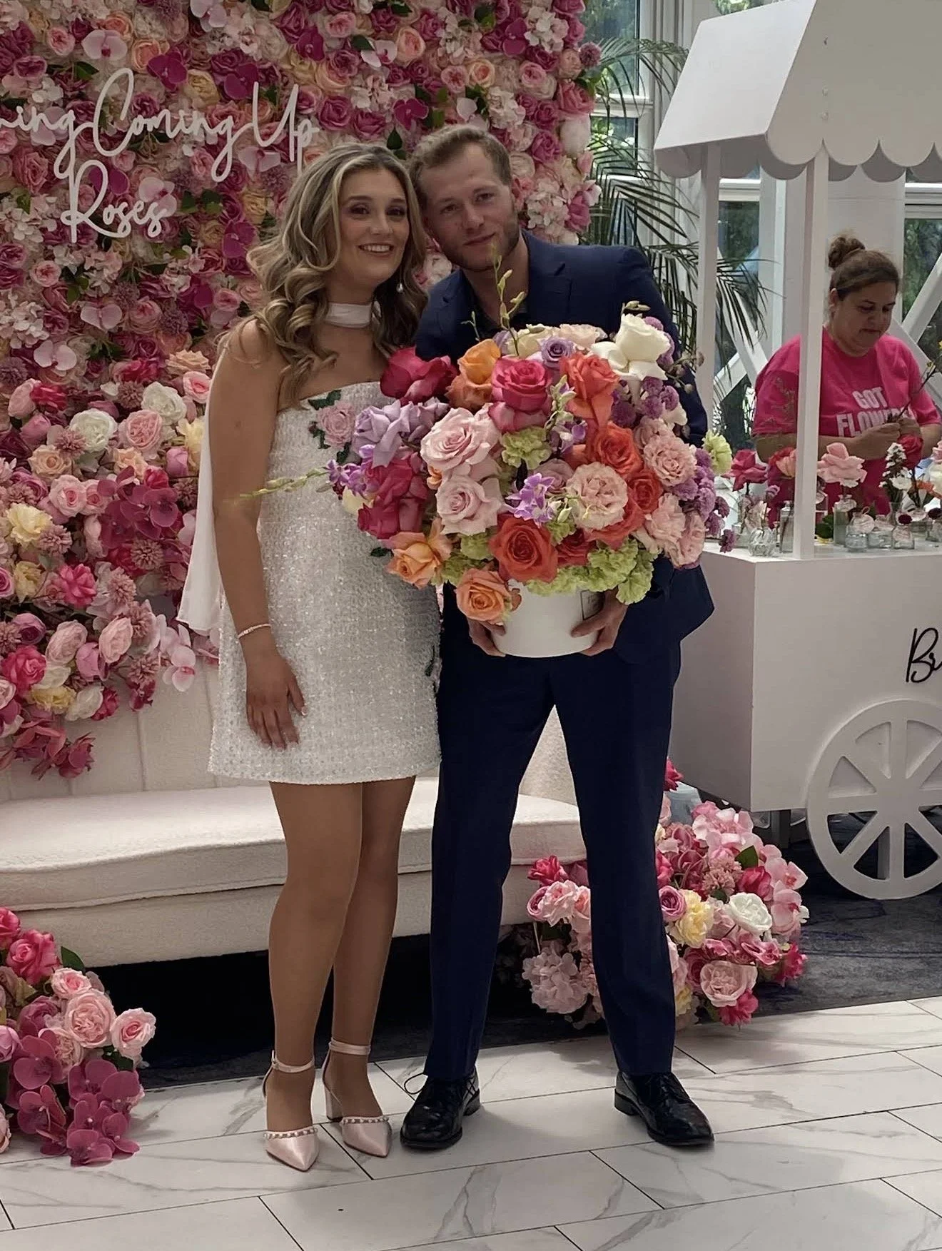 A bride and groom standing together at a wedding reception, with a backdrop of pink and white flowers and a sign that says 'Coming Up Roses.' The bride wears a white dress with floral embroidery, and the groom wears a navy suit. The groom holds a lar