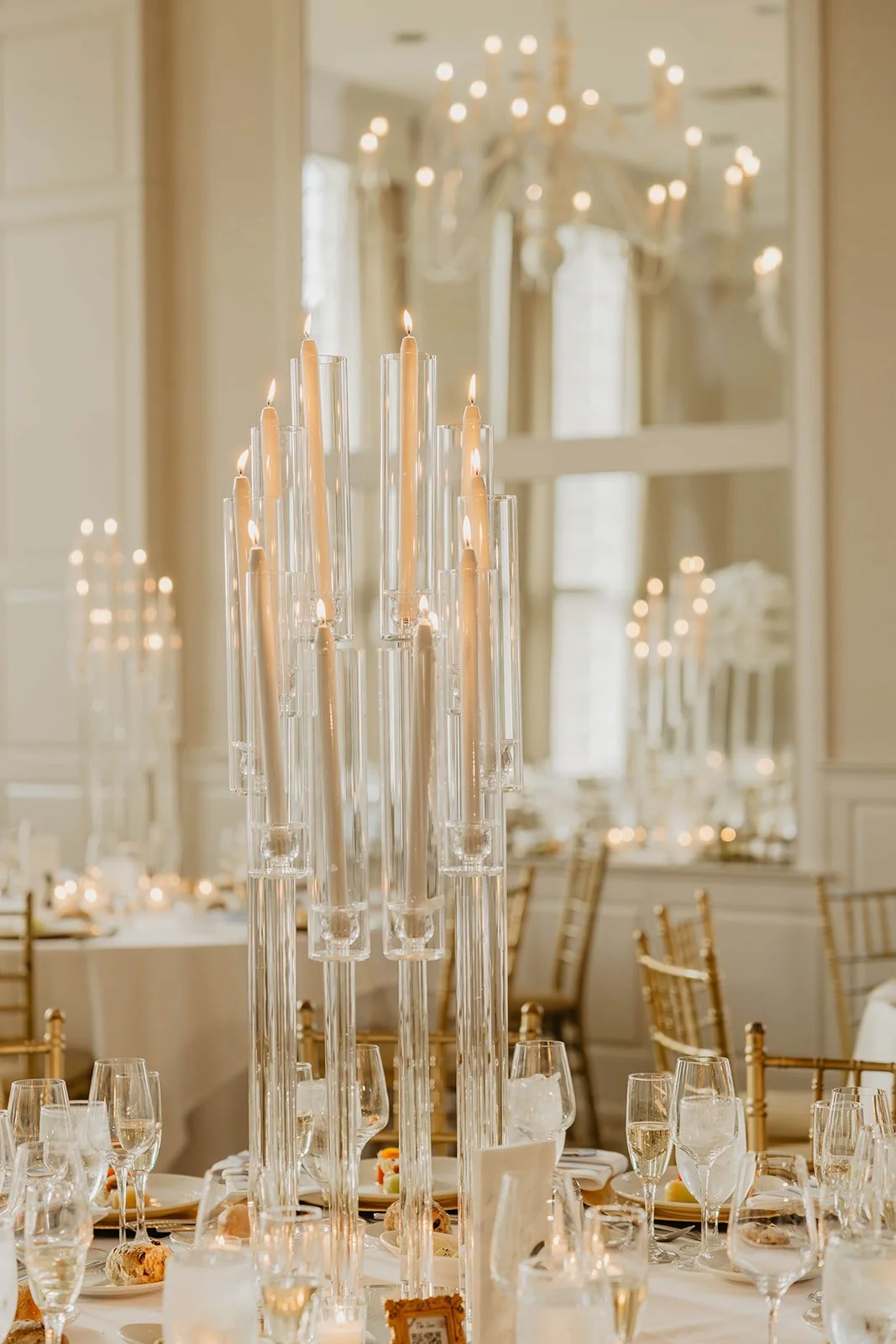 Elegant dining setup with a tall centerpiece of lit candles in glass holders, surrounded by wine glasses and plates, with gold chairs and a chandelier in the background.