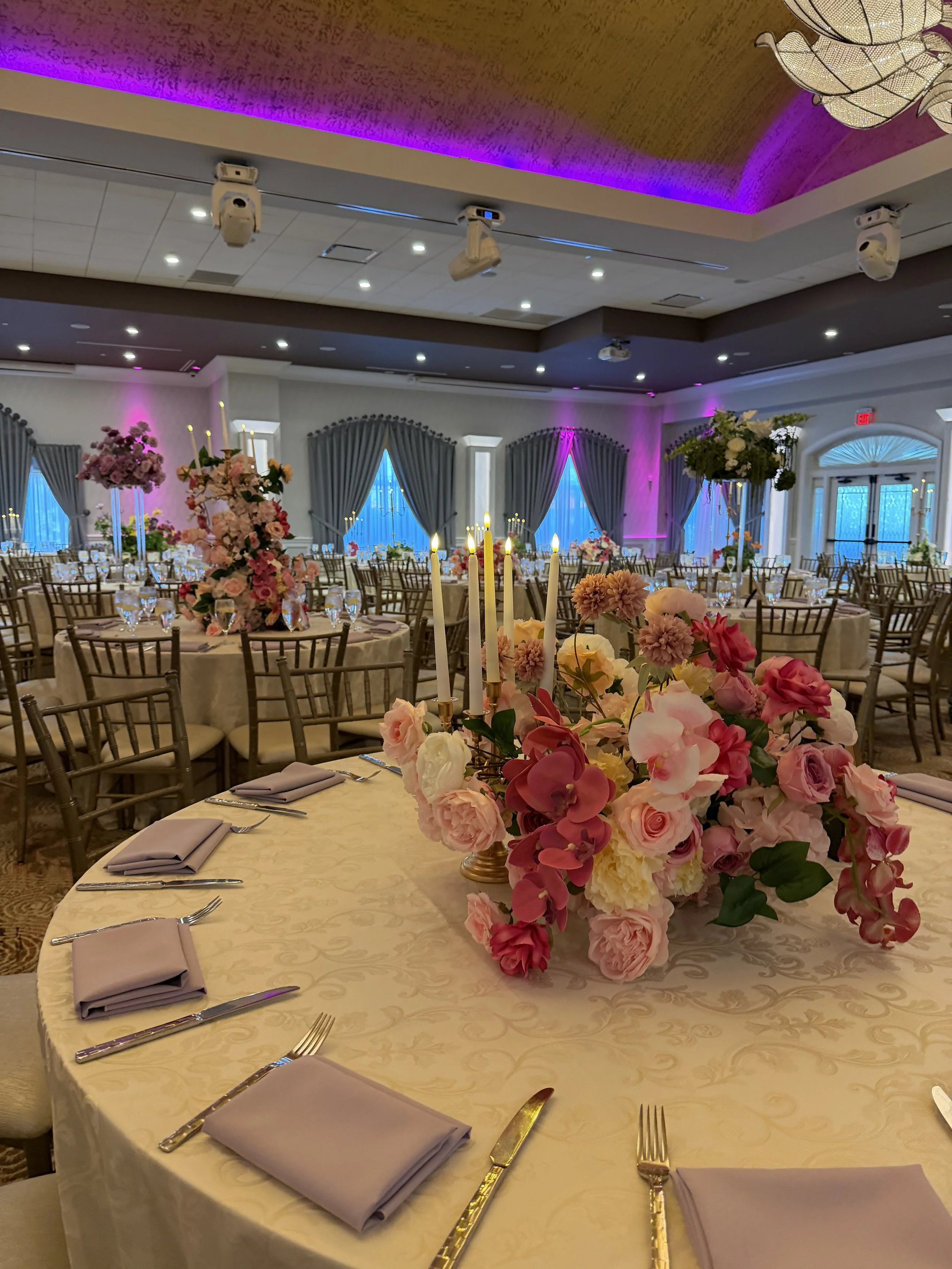 Elegant banquet hall decorated for a wedding or special event, featuring a large floral centerpiece with pink, white, and purple flowers on a round table, surrounded by chairs and set with cutlery and napkins, with draped curtains, blue lighting, and
