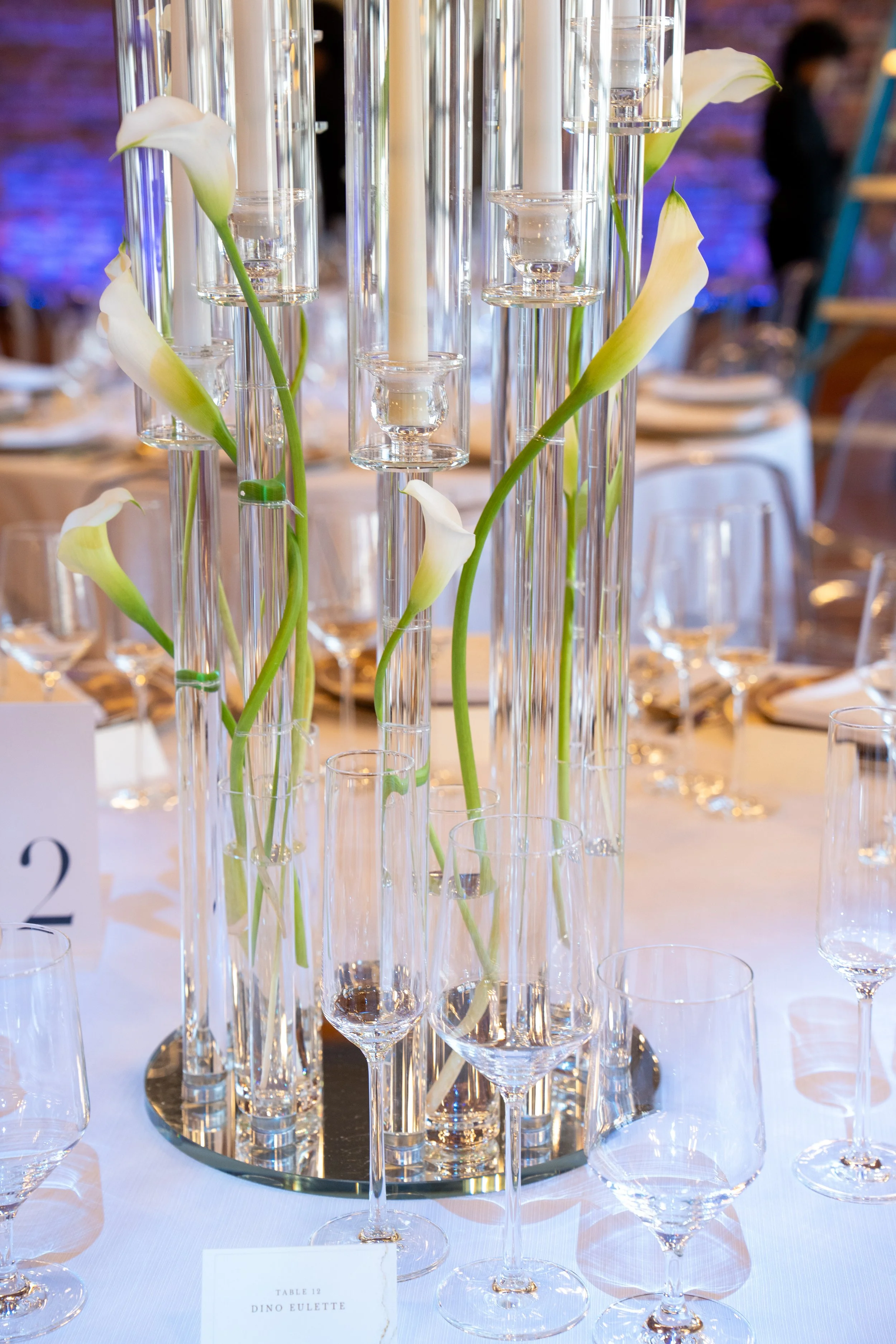 Elegant centerpiece with white calla lilies and tall glass candle holders at a decorated event table.