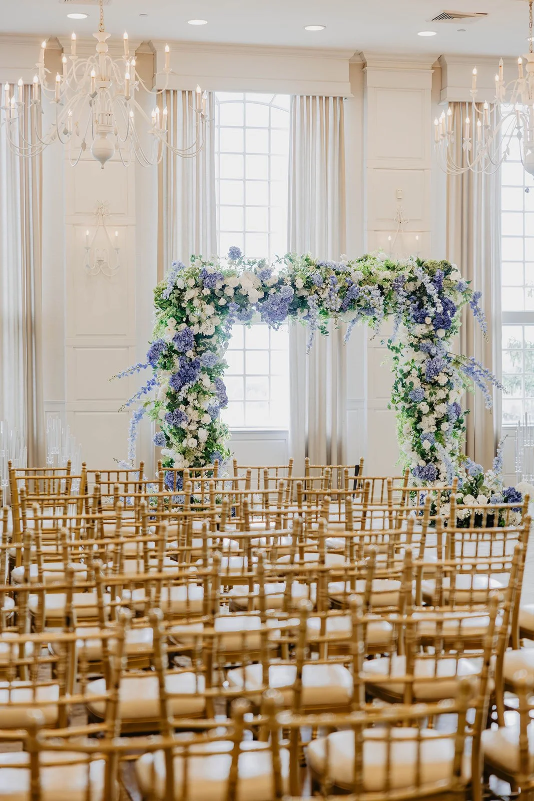 Wedding ceremony setup with rows of wooden chairs facing a floral arch in a bright, elegant room with chandeliers and large windows.