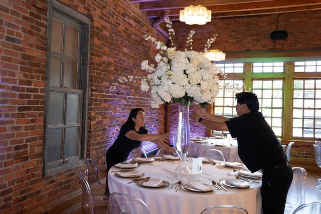 Two people arranging a large white floral centerpiece on a dining table in a restaurant with brick walls and large windows.