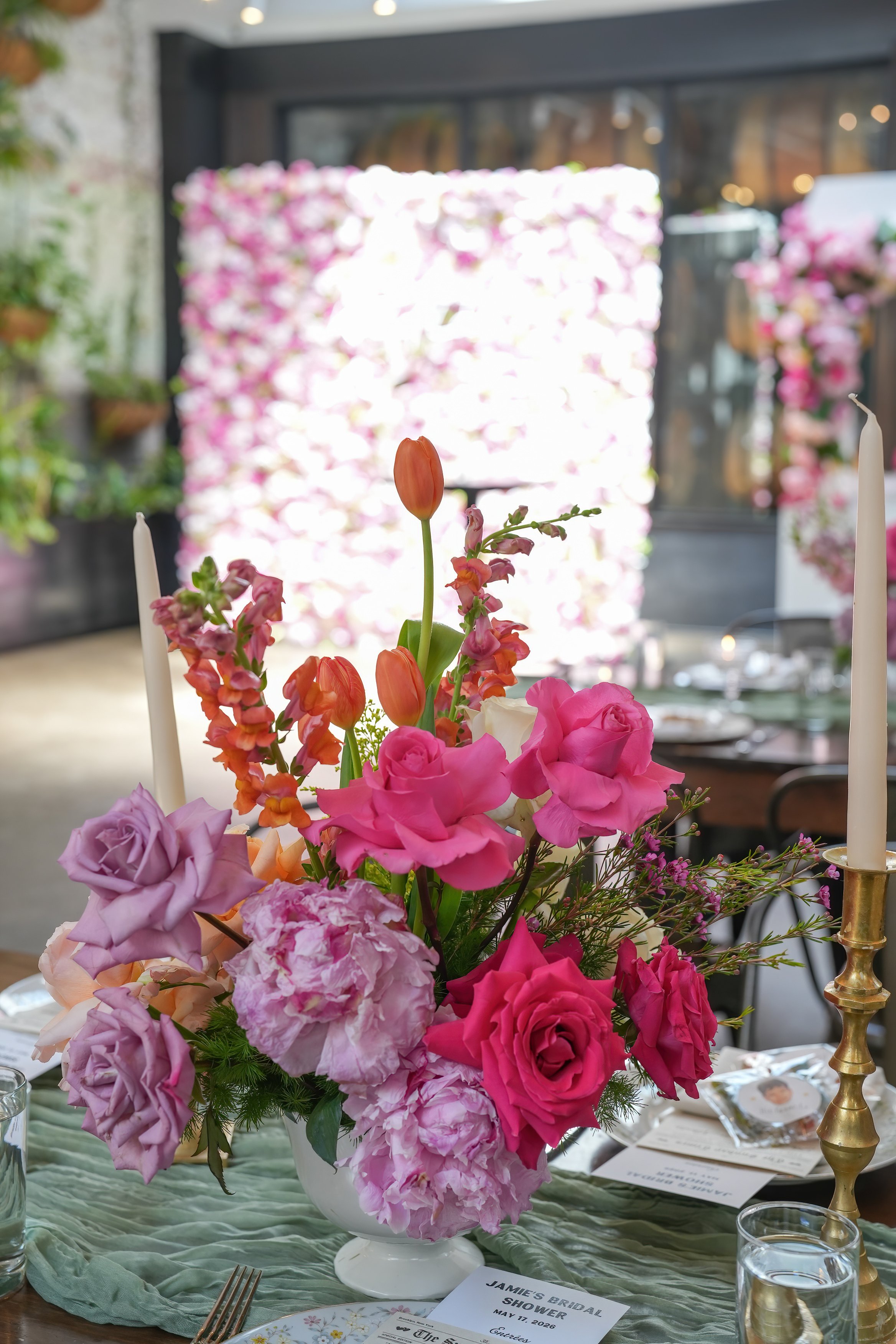 A floral centerpiece with pink, purple, and orange flowers on a table. In the background, there is a blurred floral backdrop and table setting with candles and place cards.