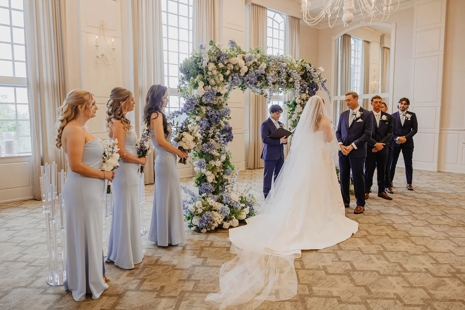A wedding ceremony with a bride and groom exchanging vows in front of an officiant, surrounded by bridesmaids and groomsmen, with a large floral arch and a chandelier in a bright, elegant room.