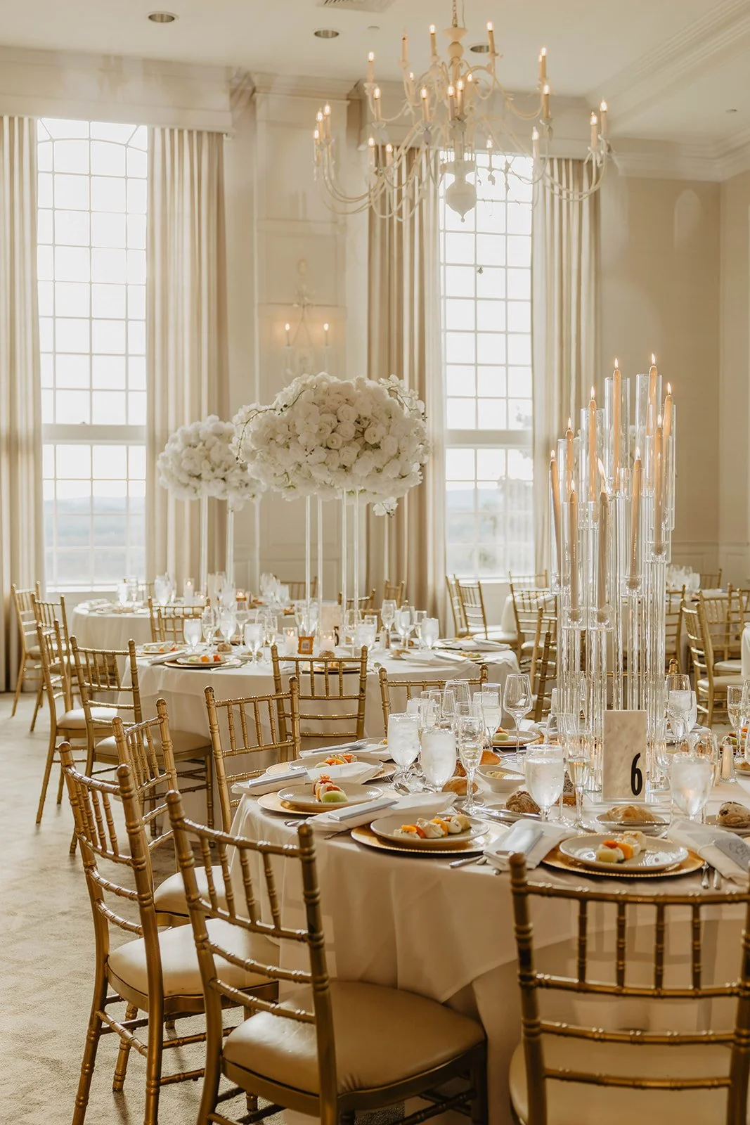 Elegant banquet hall decorated with cream-colored curtains, a chandelier, tall candle holders, and round tables with white tablecloths, floral centerpieces, and place settings.