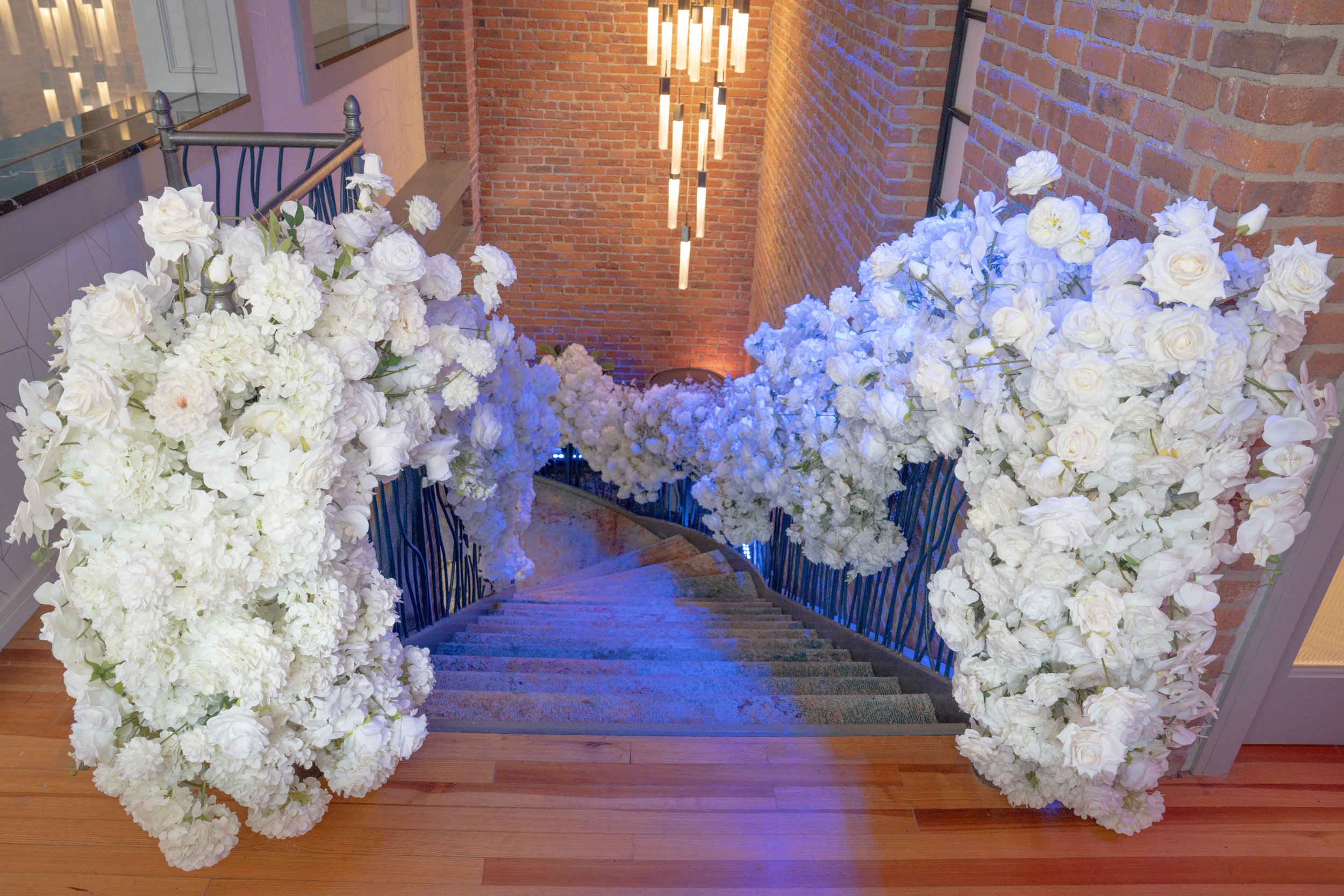 Staircase decorated with white flowers along the sides and illuminated by hanging lights on a brick wall.