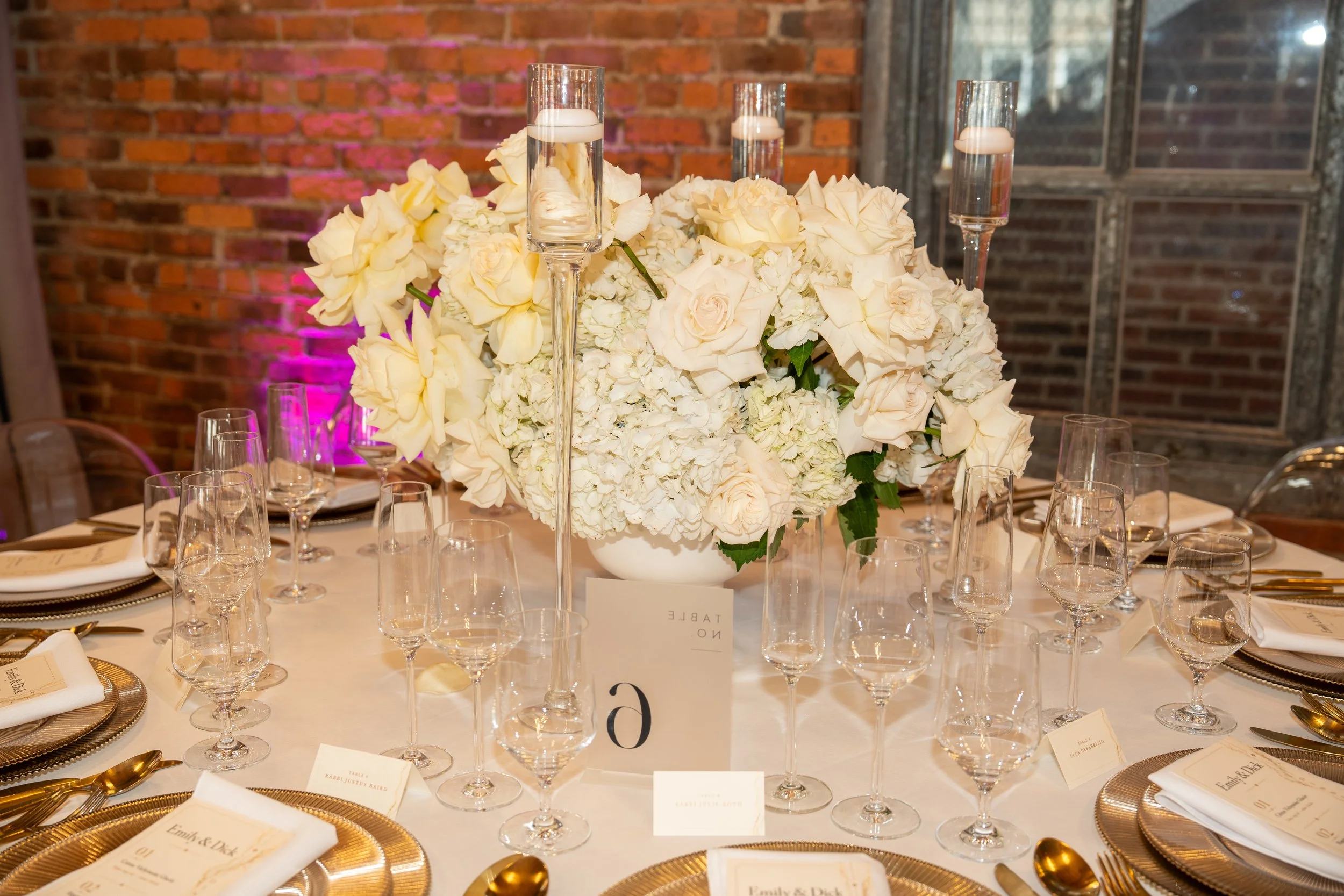 Elegant wedding reception table with a large floral centerpiece of white roses and hydrangeas, surrounded by glass candle holders, gold-rimmed plates, gold utensils, and printed place cards in front of each setting, against a brick wall backdrop with