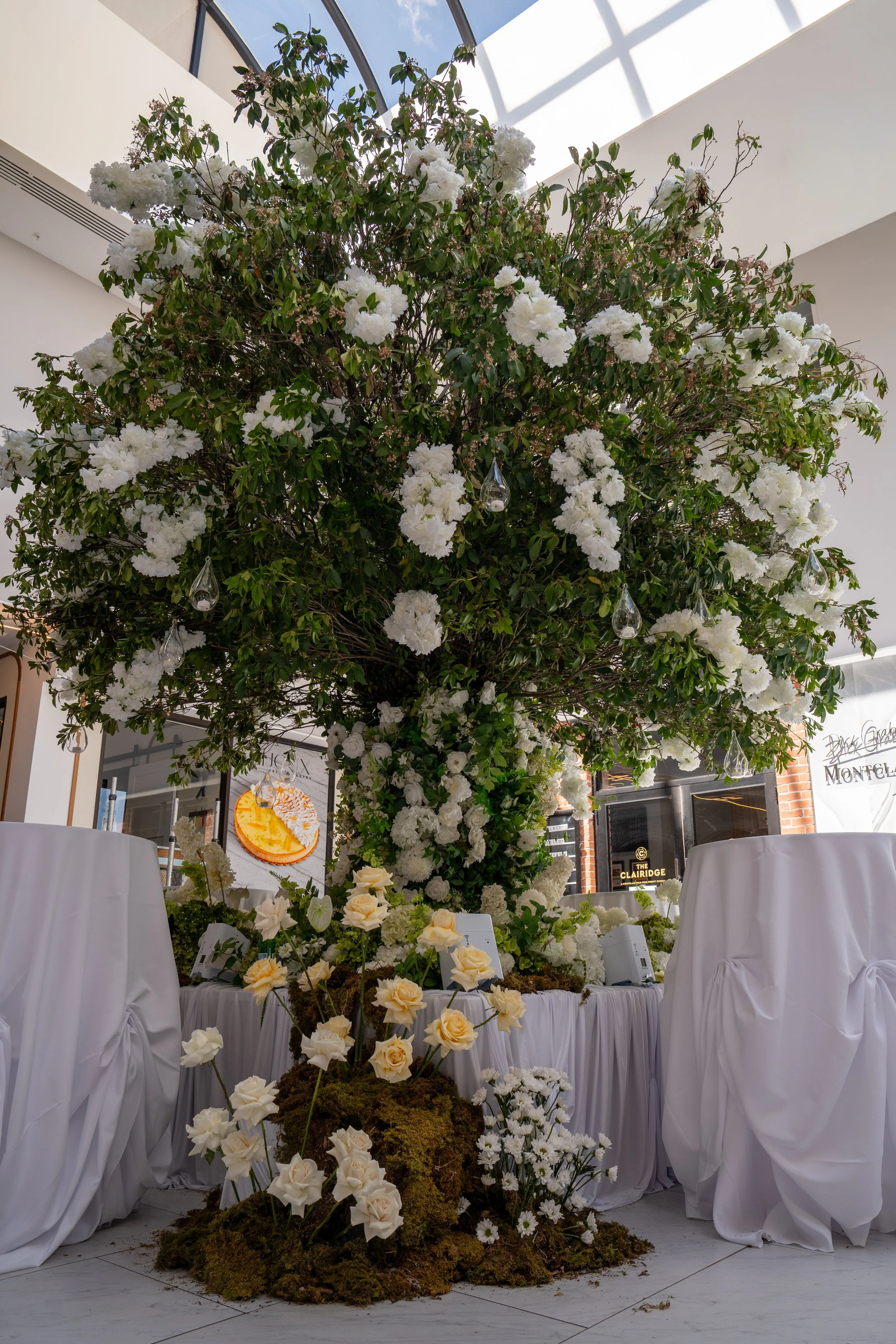 Floral display with white flowers and greenery on tables and a large blooming tree inside a building with a glass ceiling.