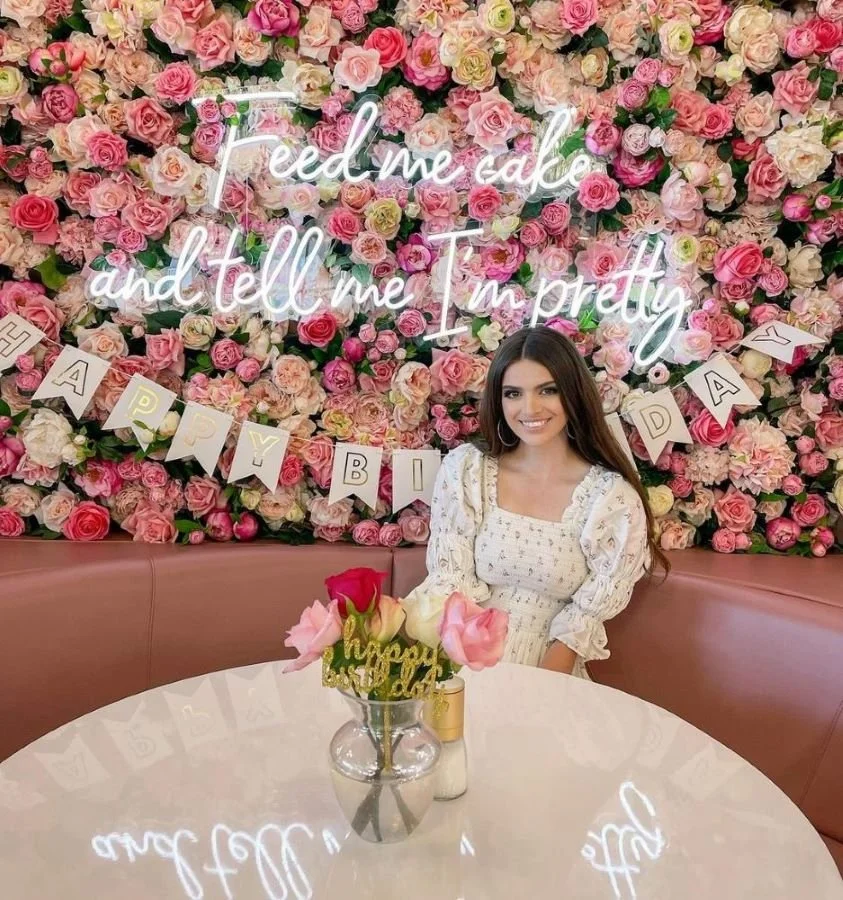 A woman sitting at a table with a bouquet of pink and white roses with a "Happy Birthday" sign in a glass vase. Behind her is a wall decorated with pink, white, and cream roses and a neon sign that reads, "feed me cake and tell me I'm pretty." There is also a banner that spells out "HAPPY BDAY." She is smiling and wearing a white dress with puffed sleeves.