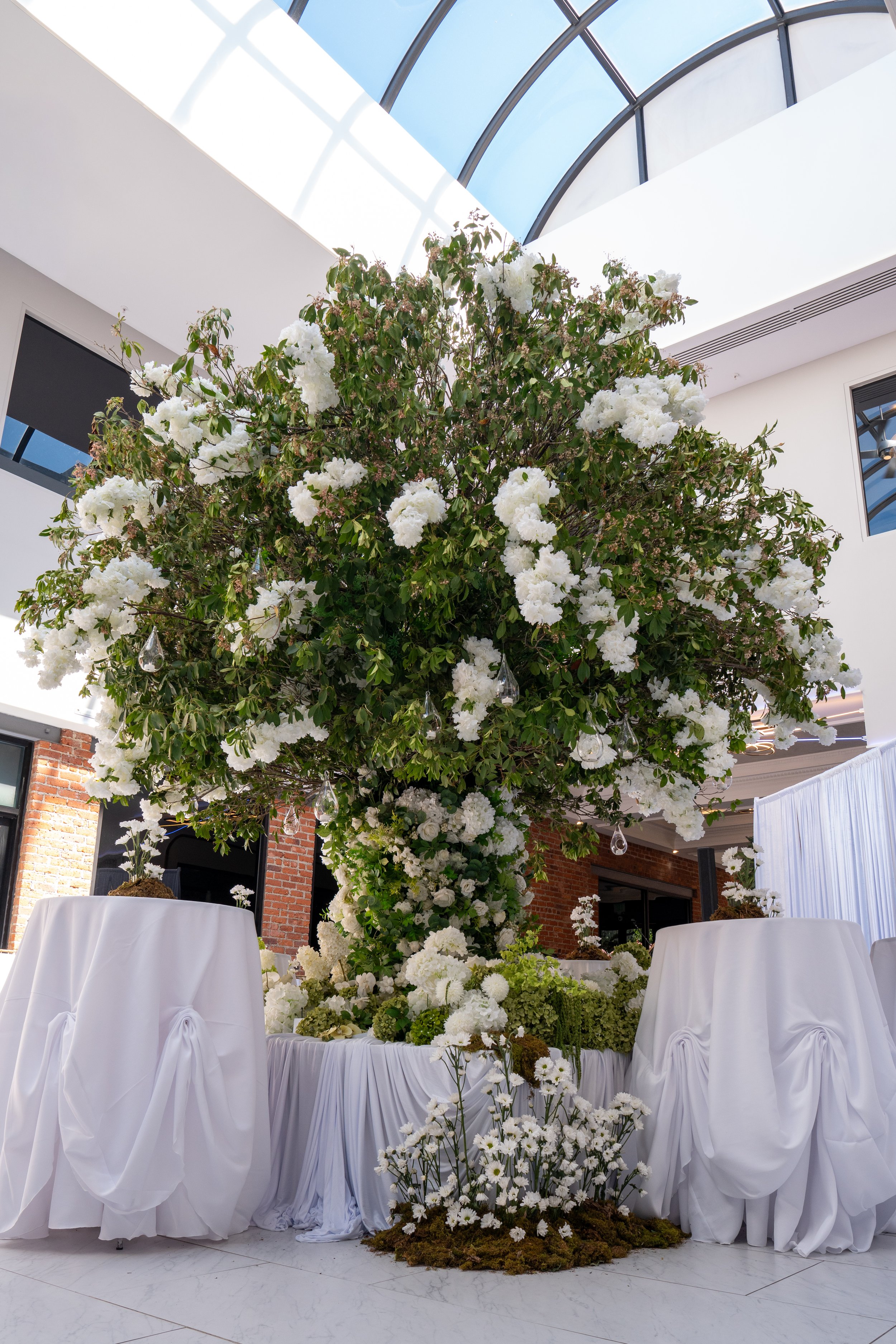 A large white floral arrangement with white flowers and greenery, set on a draped table, in a modern indoor space with a glass ceiling and brick walls.