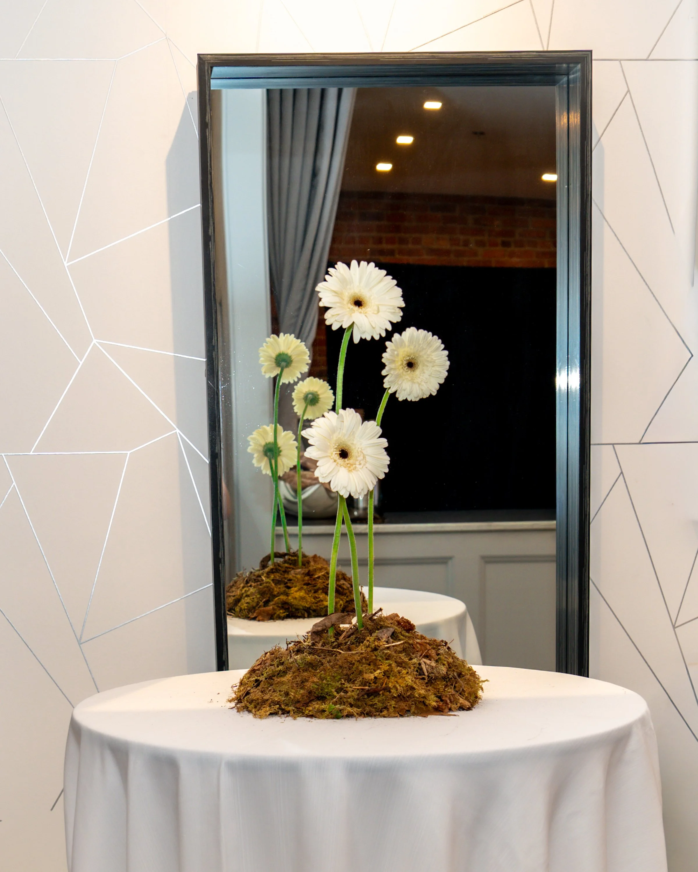 A potted plant with white flowers on a table, with a mirror and a geometric-patterned wall in the background.