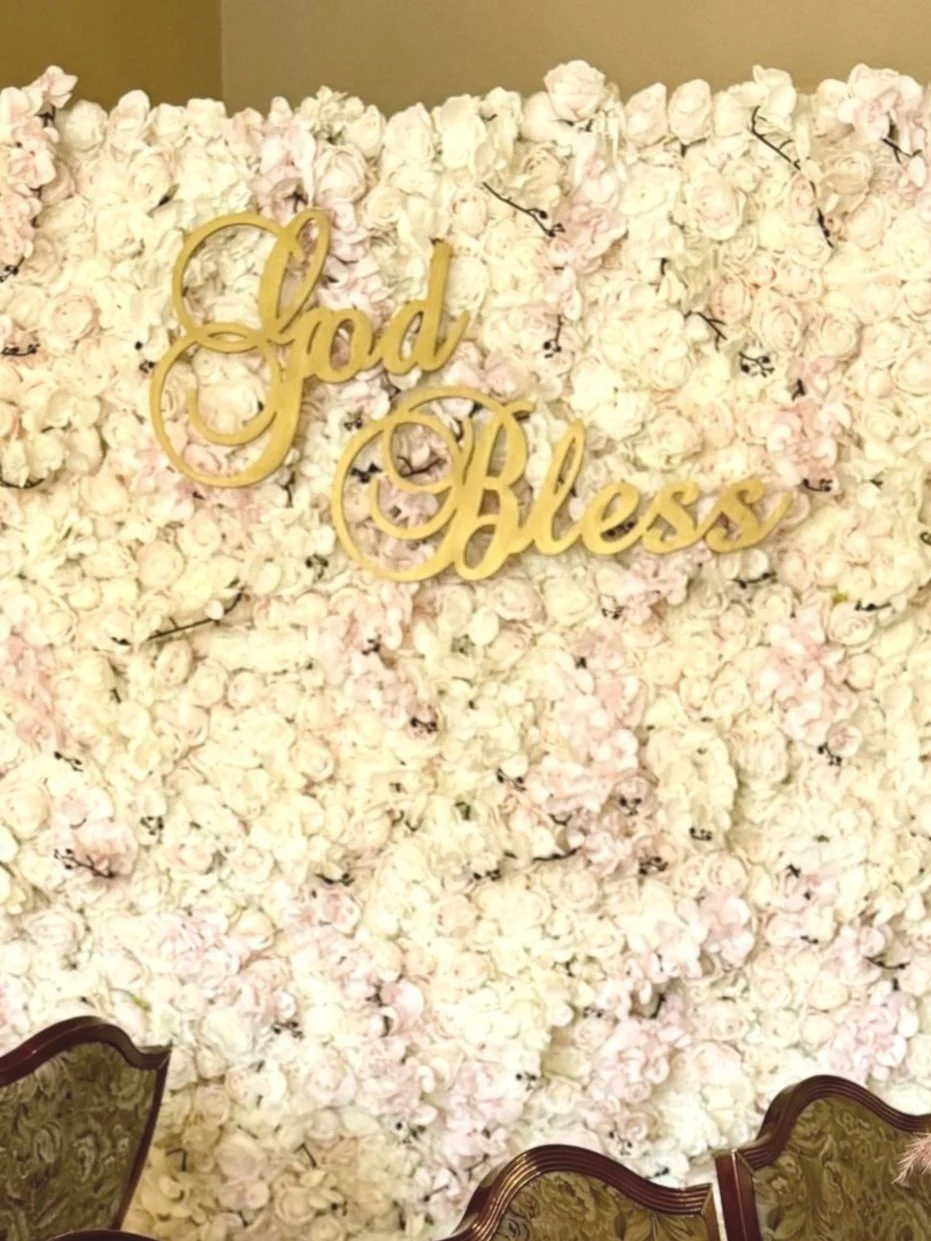 A decorative wall made of light pink and white roses with a gold cursive sign that reads 'Good Bless' in the center. flower wall rental in nj permanent installations flower murals, bloom bars, audio guest book, wedding flowers photo booth