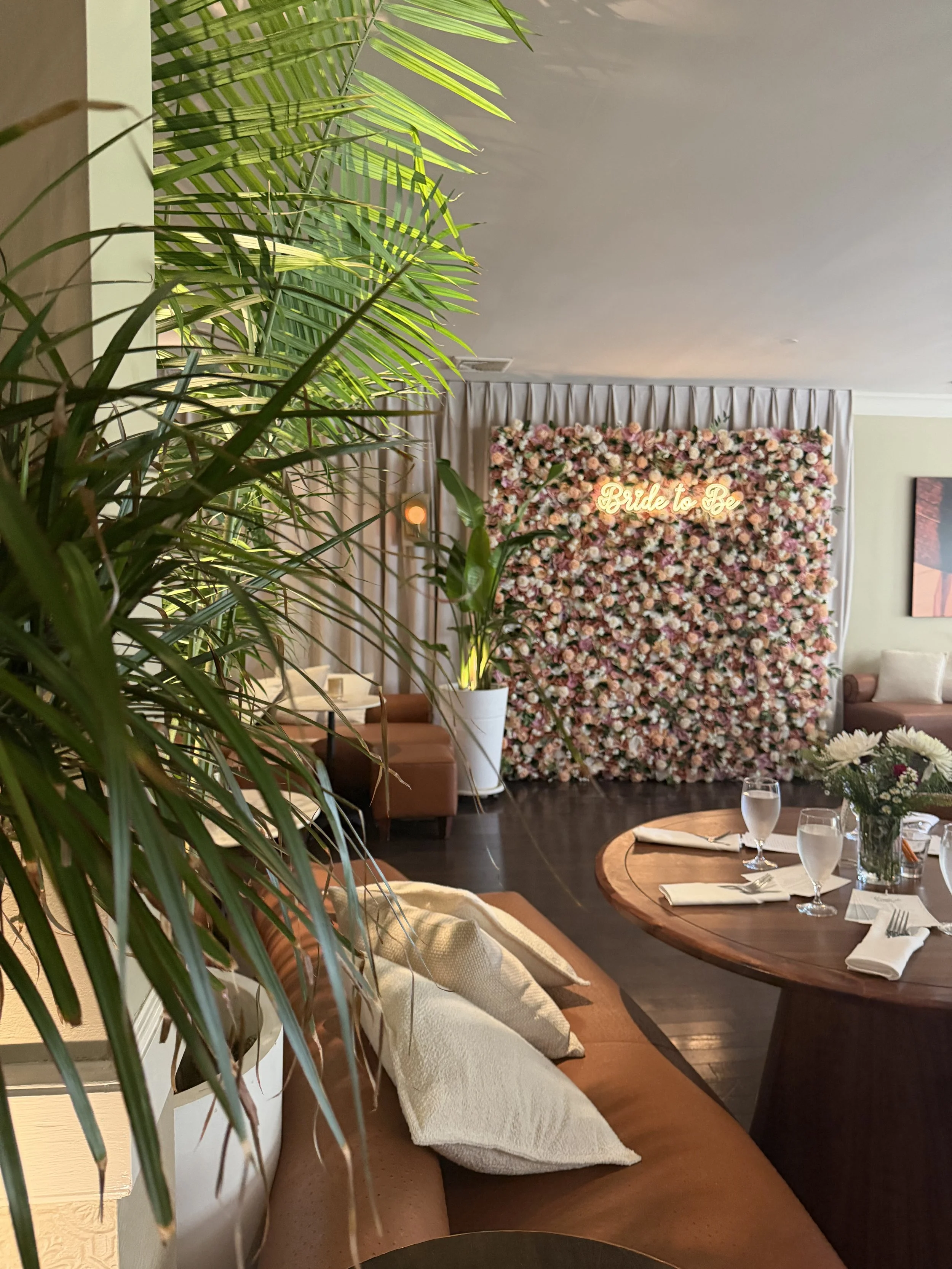 Indoor dining area decorated for a bridal event, featuring a large floral backdrop with a neon sign that reads 'Bride to Be,' surrounded by tables with place settings and a vase of white flowers, with green plants in the foreground.