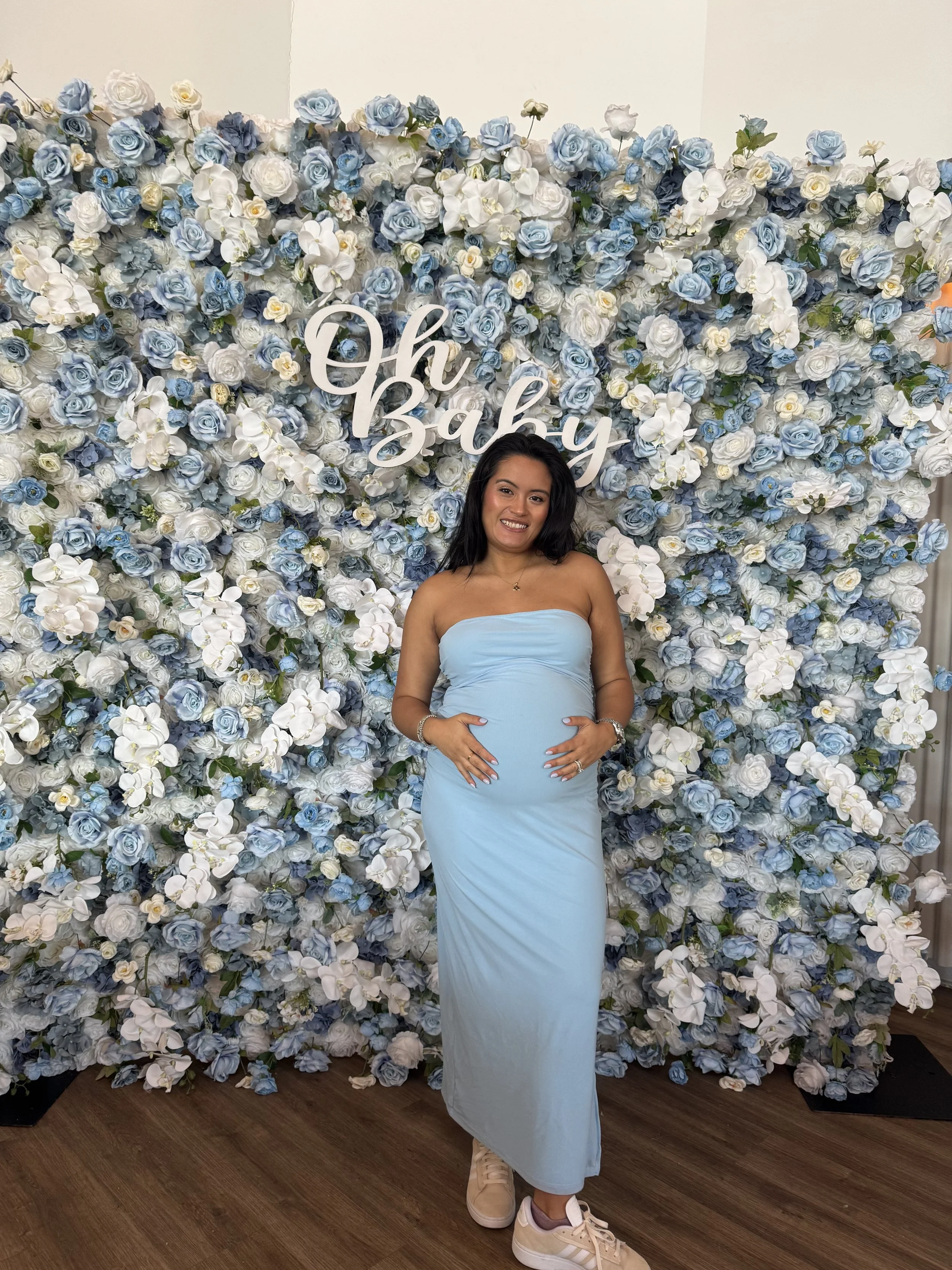 A woman in a strapless, light blue dress standing in front of a floral backdrop of white and blue roses with a sign that reads "Oh Baby." flower wall rental in nj permanent installations flower murals, bloom bars, audio guest book, wedding flowers