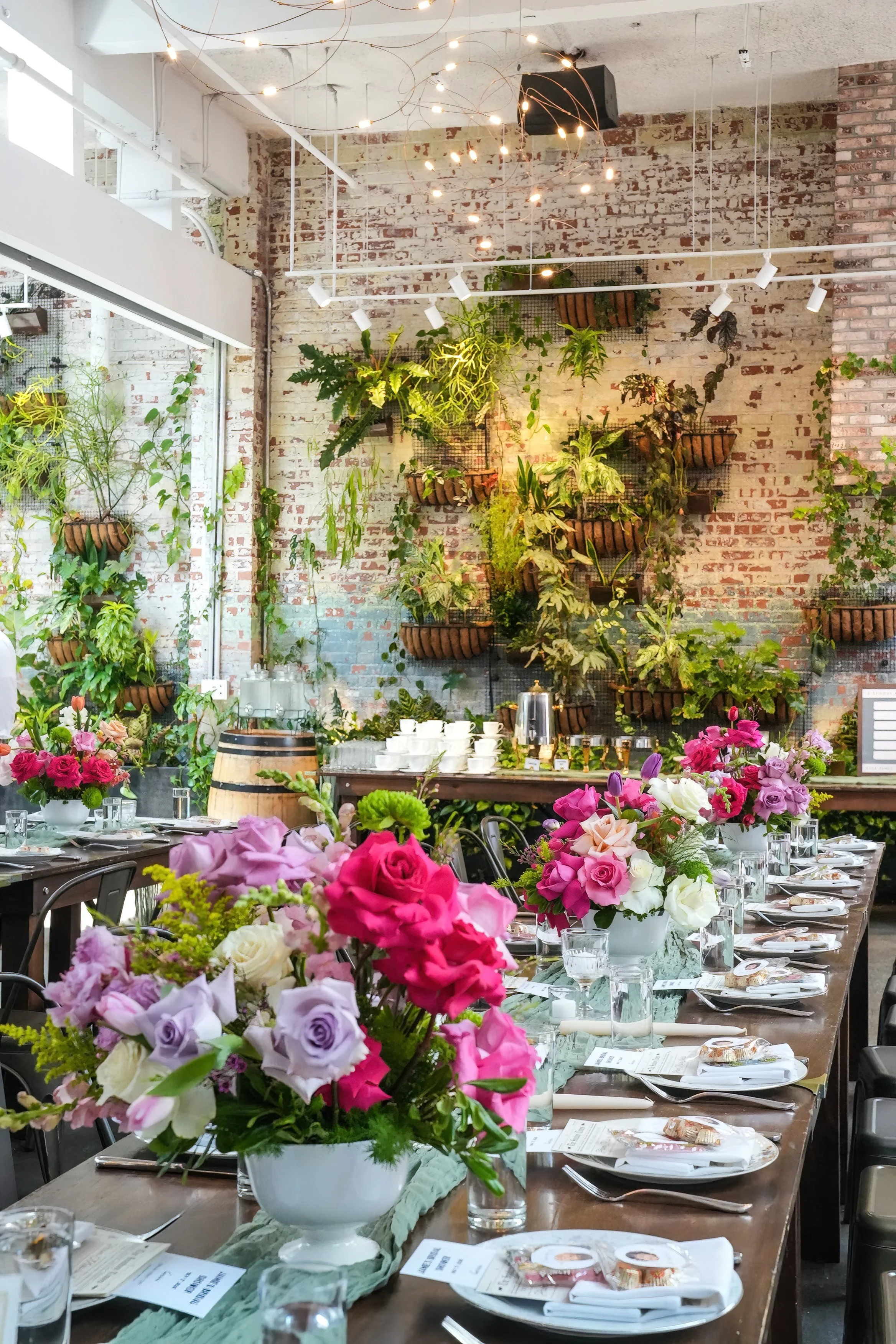 A banquet table decorated with colorful flower arrangements, set for a formal event in an industrial-style venue with brick walls and hanging plants.