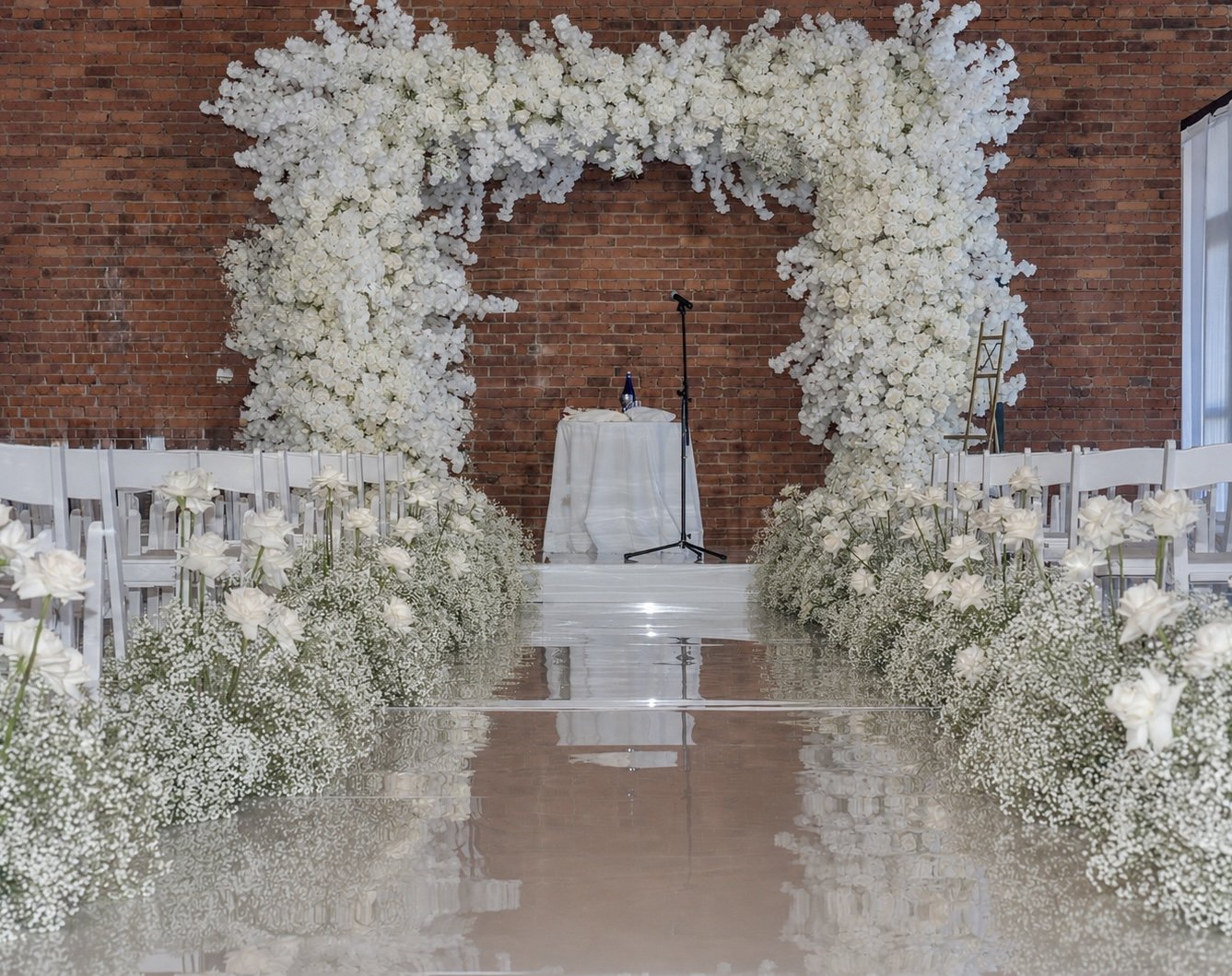 Wedding ceremony setup with a large white floral arch, white chairs lined up on each side, a small table with a microphone stand in front of a brick wall.