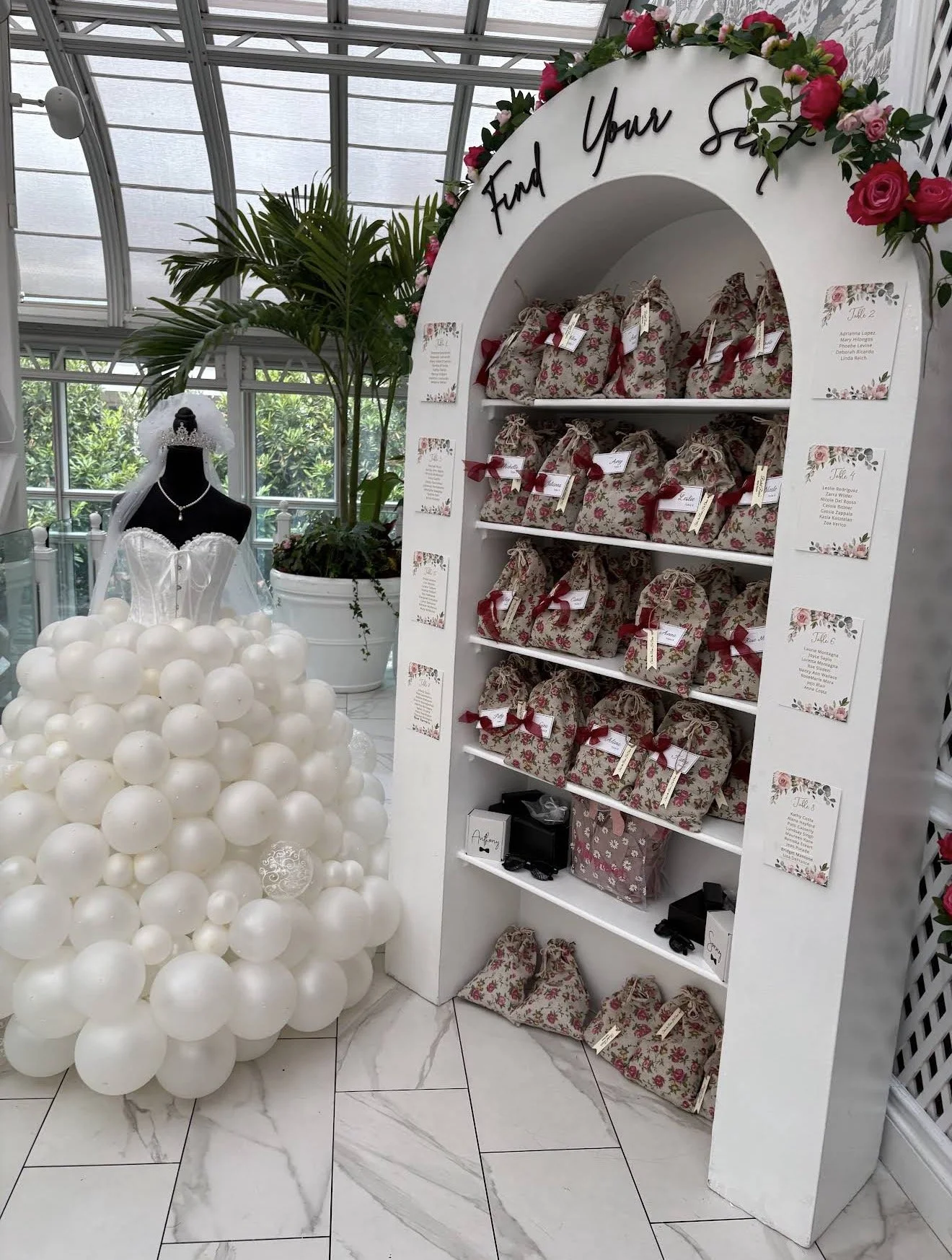 A display with a white arch that says 'Find Your Seat' decorated with pink and red flowers. Inside the arch are decorated gift bags with floral patterns and red ribbons, each labeled with a name. To the left, there is a mannequin dressed as a bride w