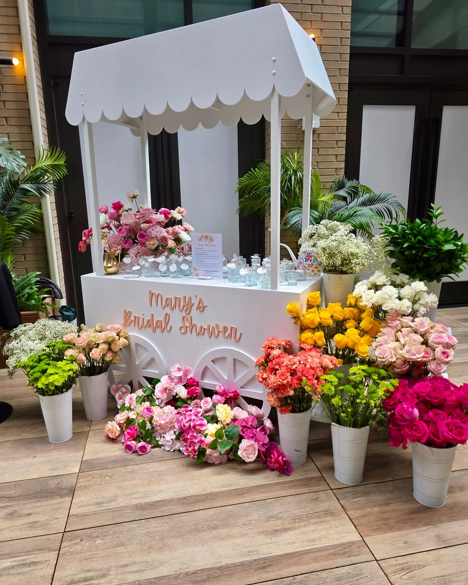 A decorative cart with a white canopy and pink, yellow, and white flowers arranged around it, with a sign reading 'Mary's Bridal Shower' on the front.