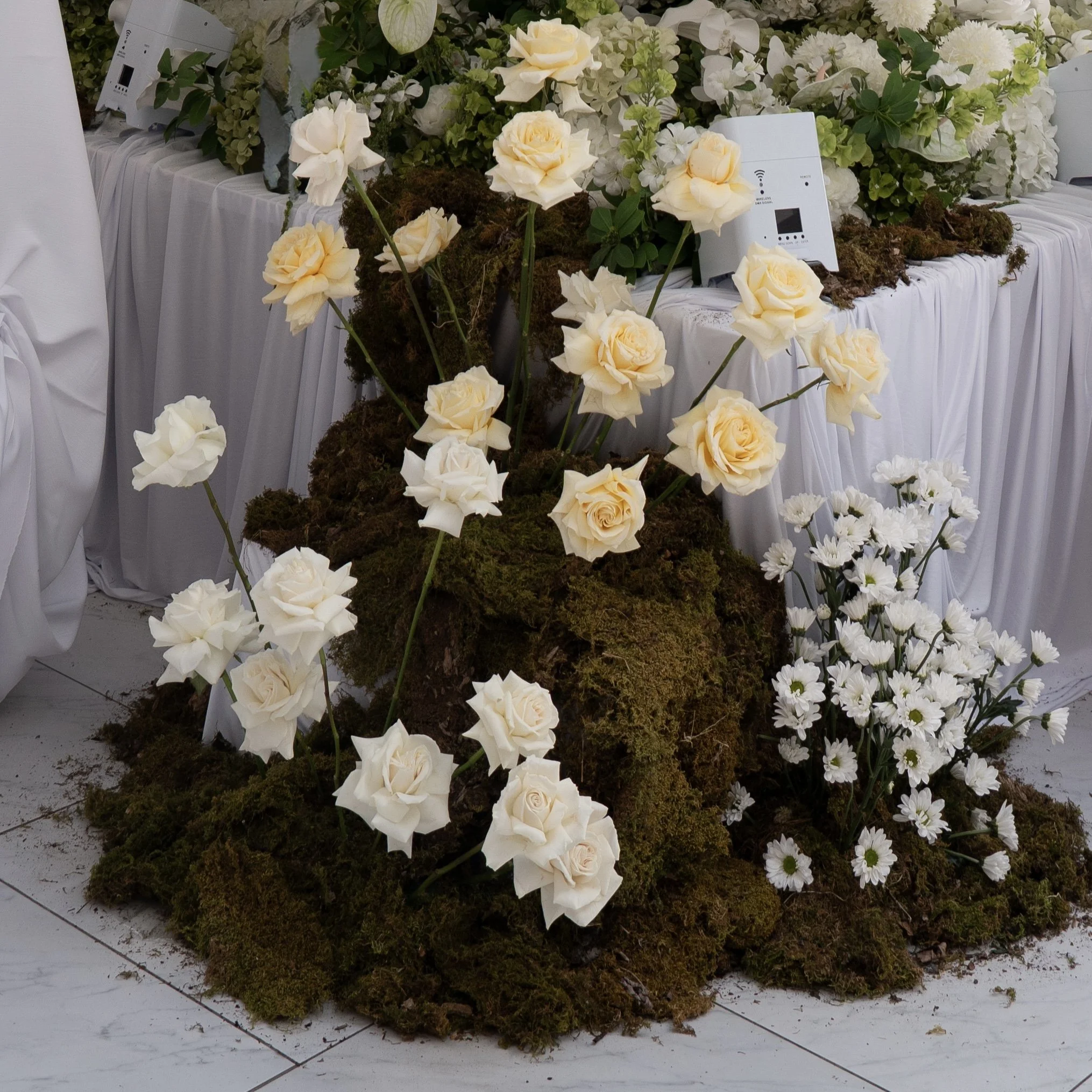 A floral arrangement with white and cream-colored roses, white daisies, and green foliage on a white table decorated with cloth, featuring A/C units and electronics among the flowers.