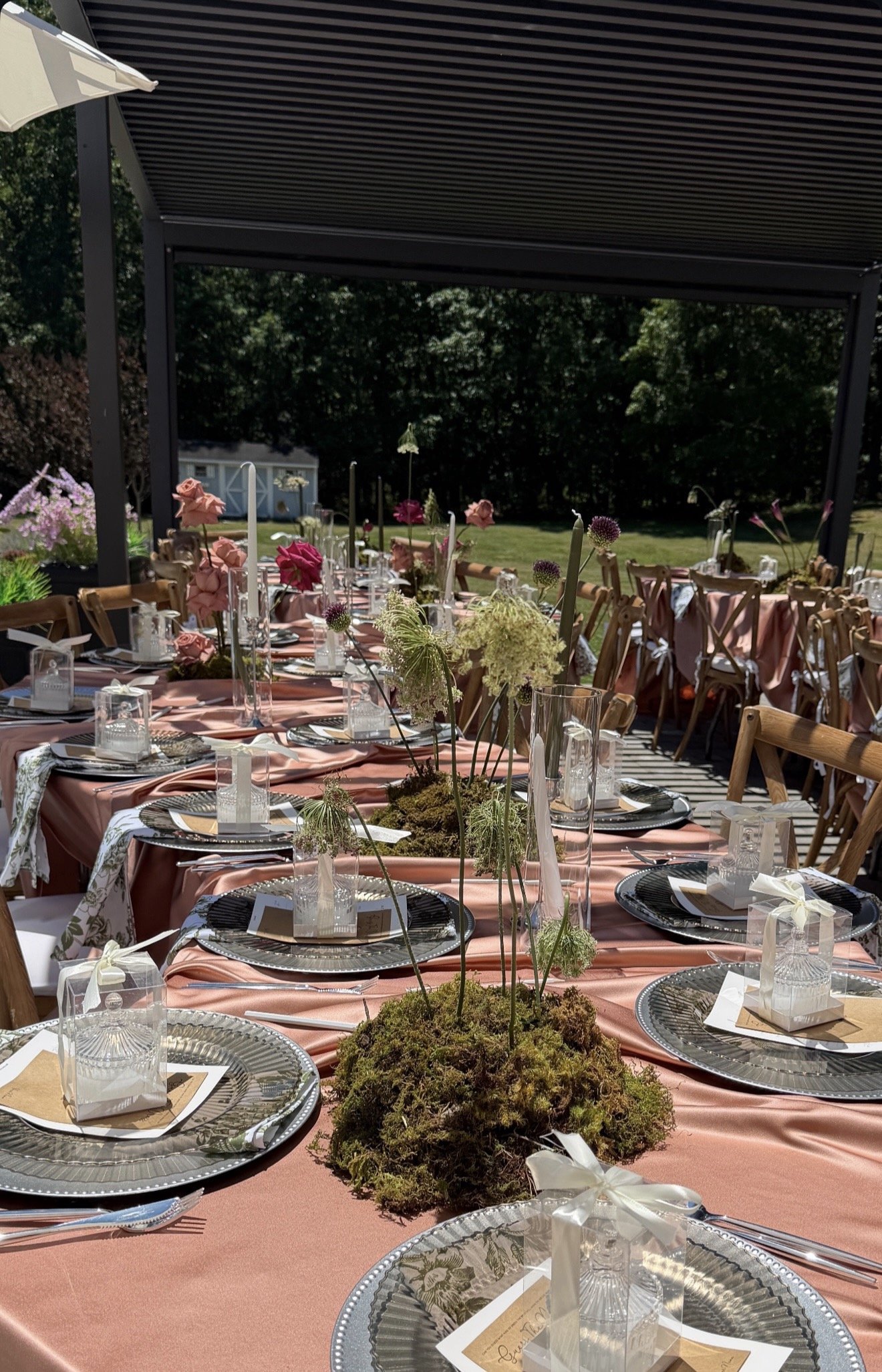 Outdoor dining setup with long tables covered in pink tablecloths, decorated with floral centerpieces, set with plates, napkins, glasses, and silverware under a large black pergola, with a grassy yard and trees in the background.
