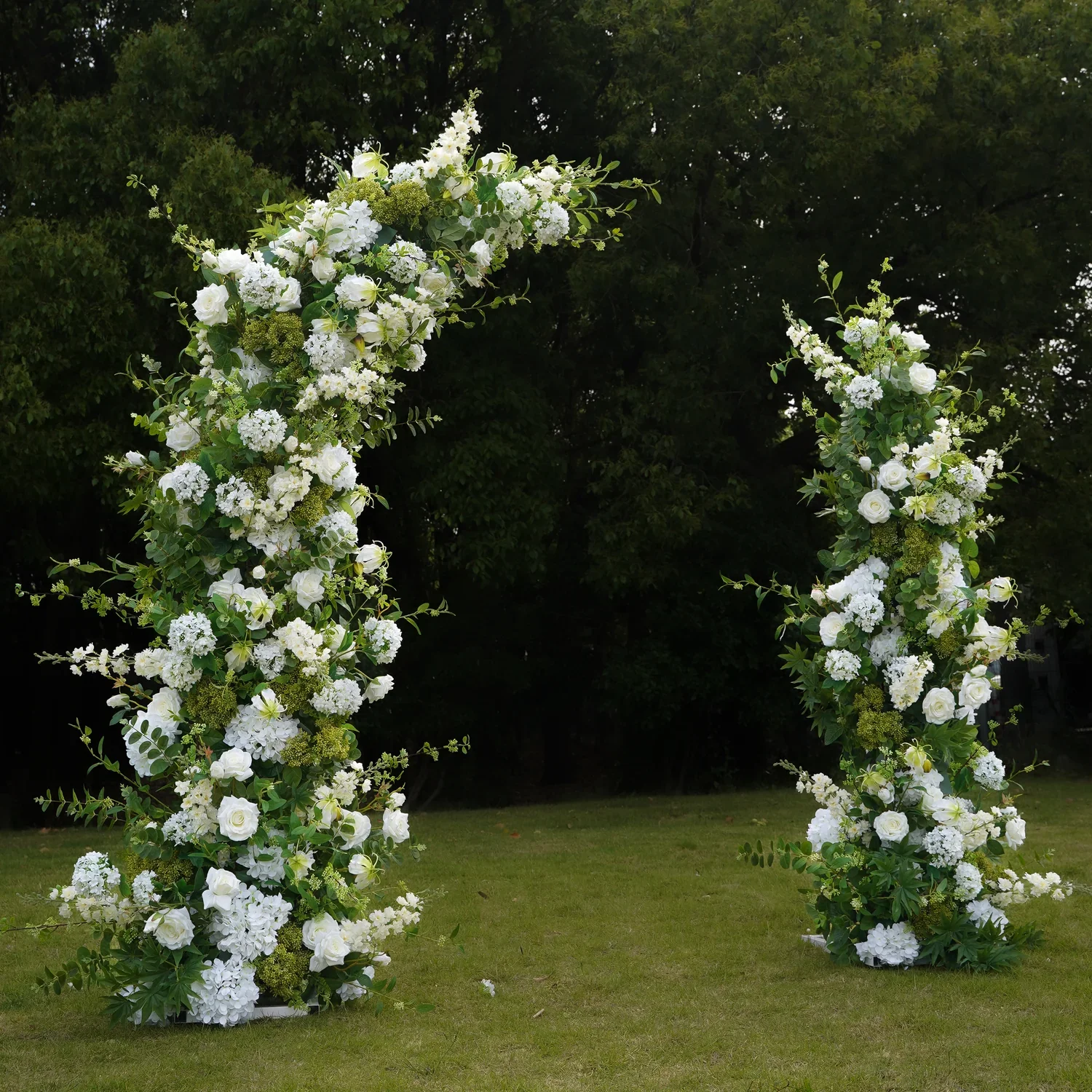 White floral arch with roses, hydrangeas, and greenery on a grassy lawn with trees in the background. wedding arch best