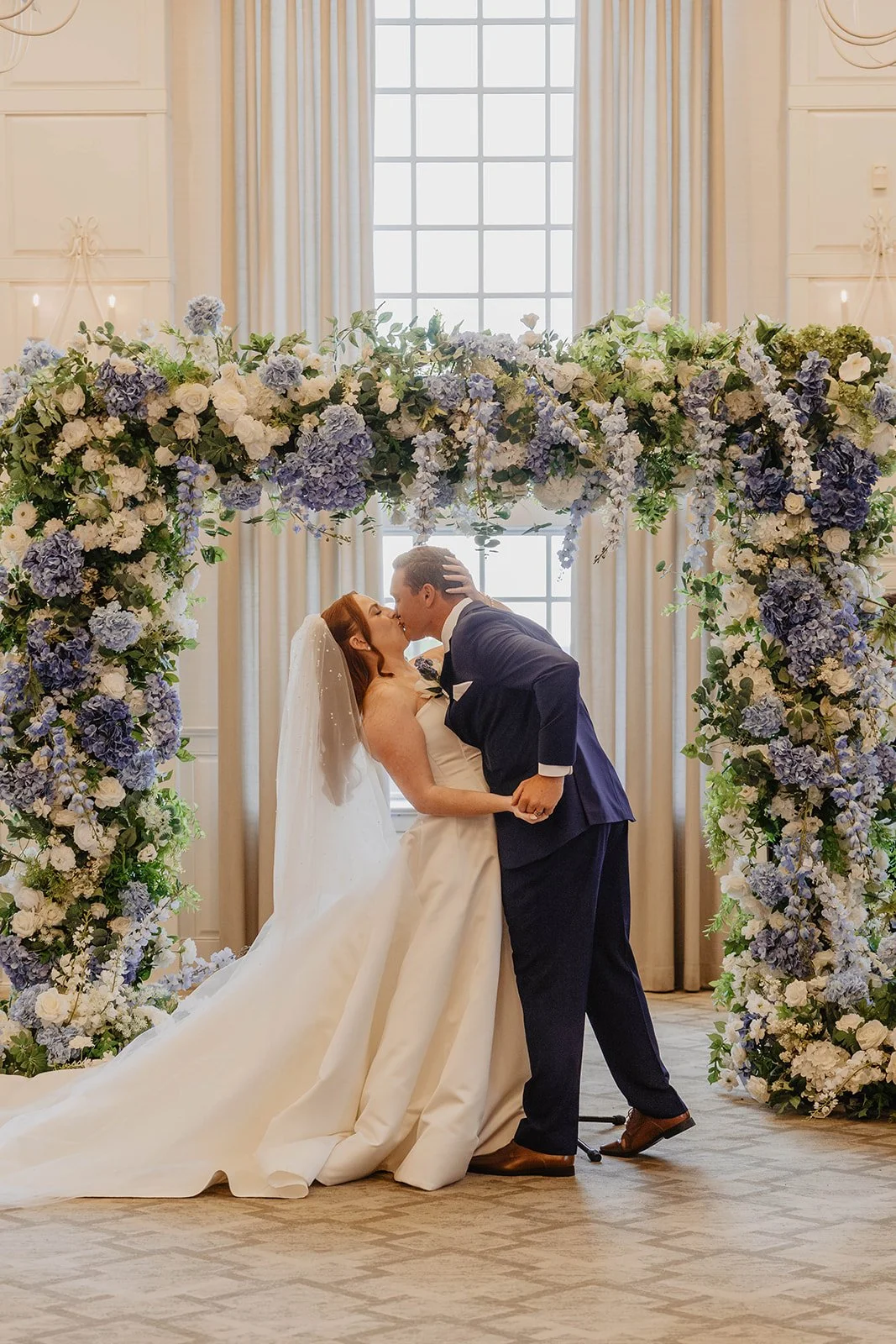 A newlywed couple sharing a kiss under a floral wedding arch in a bright room with large windows and cream-colored curtains.