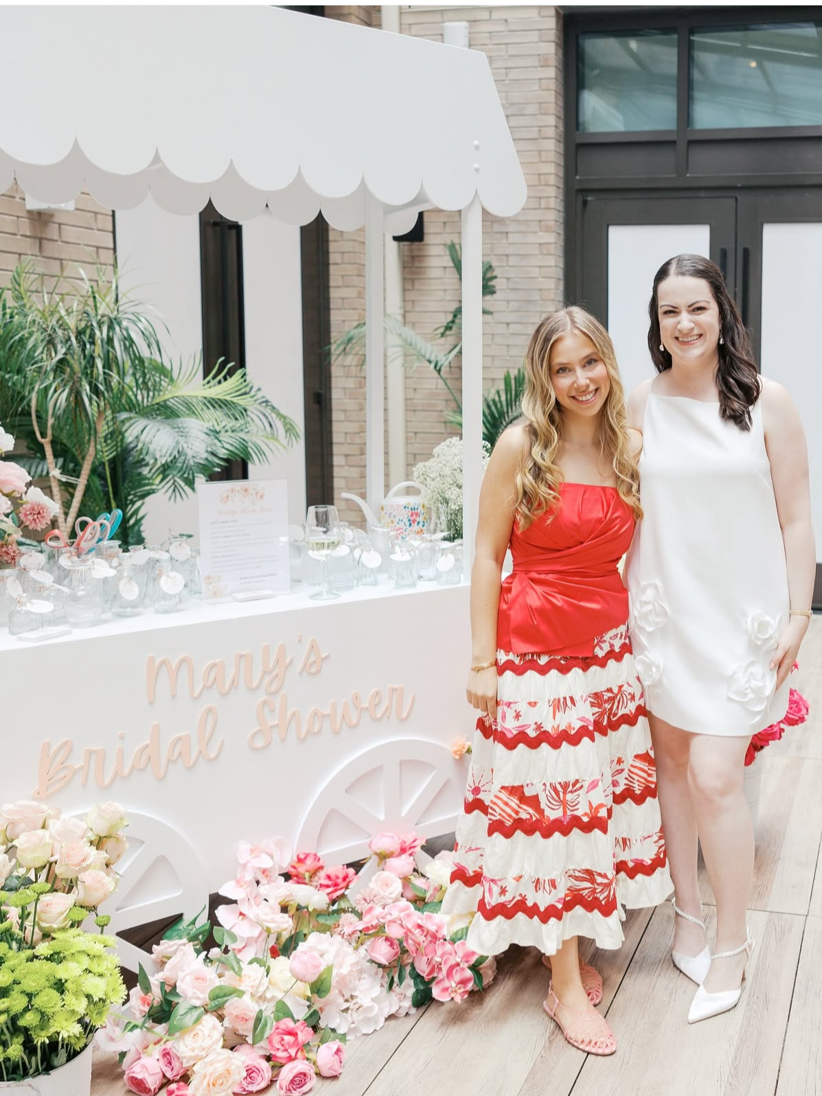 Two women standing next to each other at a bridal shower, with a decorated table behind them that has flowers, drinks, and a sign reading 'Mary's Bridal Shower', set against a modern building background.
