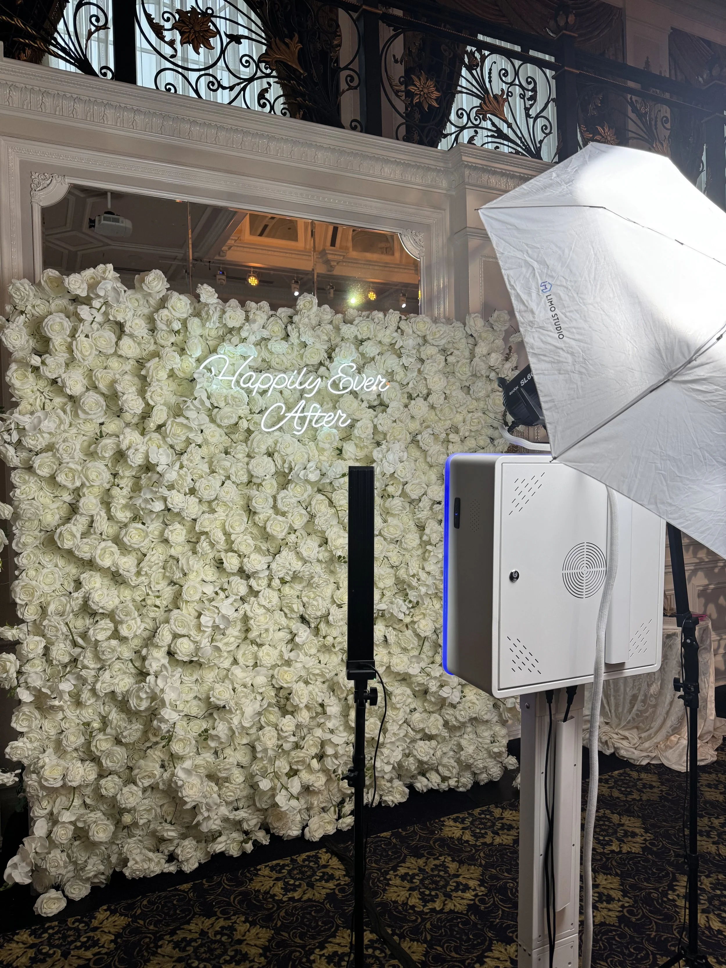 A photo booth setup at a wedding reception with a backdrop of white roses and a neon sign that reads 'Happily Ever After'. There is studio lighting equipment and a large umbrella in front of the backdrop. best photo booth with flower wall in nj ny