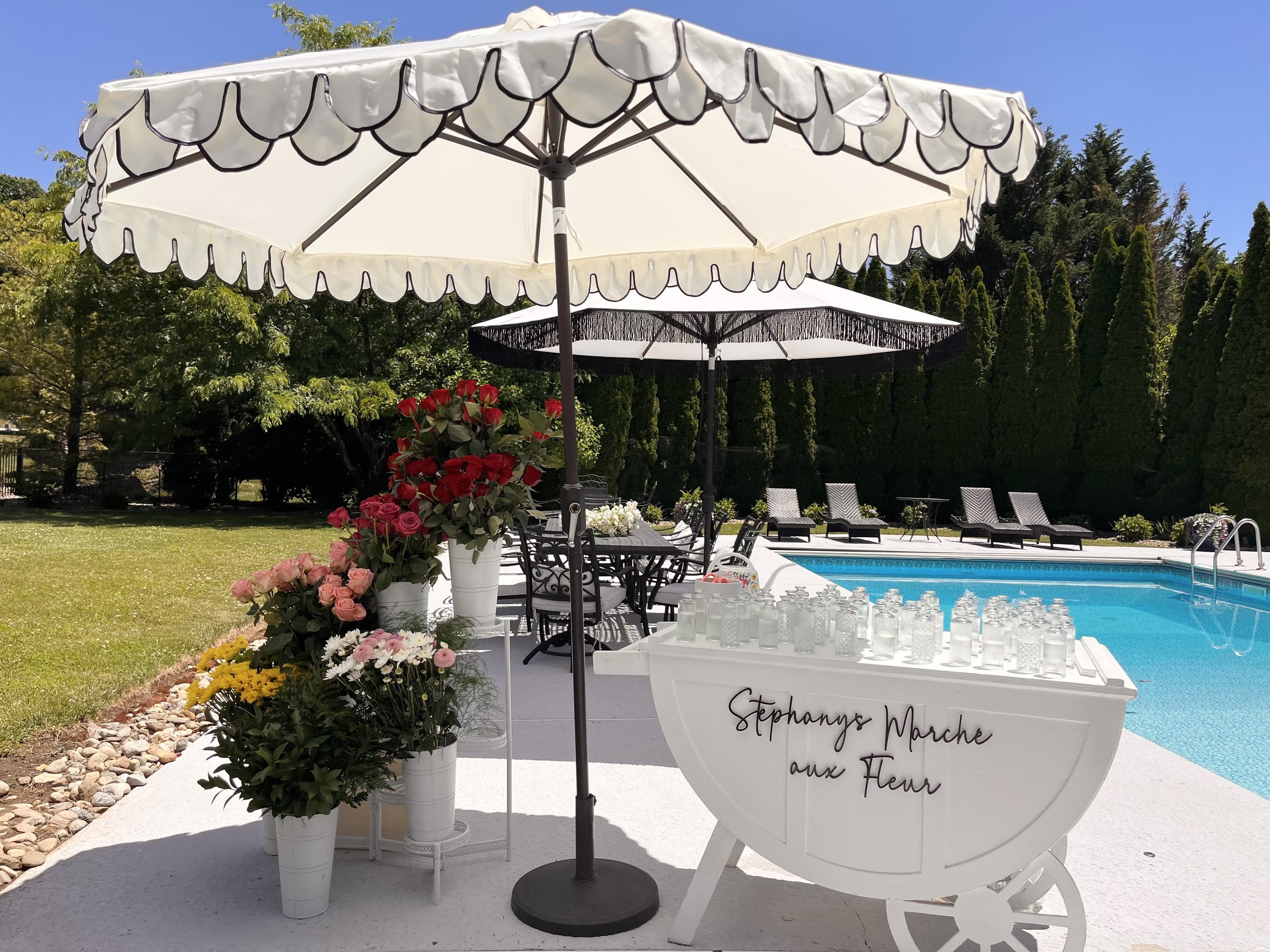 Outdoor setting with a white boat-shaped sign that reads "Stephanys Marche aux Fleurs" next to a swimming pool. There are several white planters with pink roses and yellow flowers, and two large white patio umbrellas providing shade. Outdoors, with t