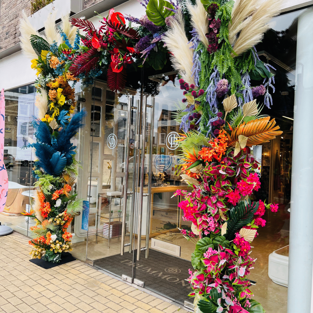 Colorful floral arch with various flowers and greenery decorating the entrance of a building with glass doors.