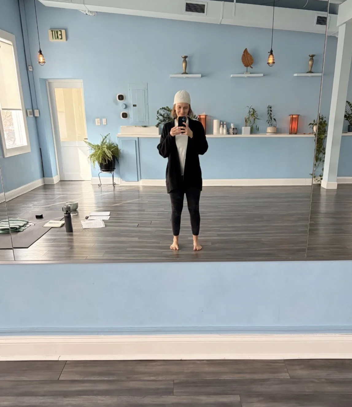 Halli Lou Faulkner taking a photo of herself standing in a big, wall mirror in a powder blue yoga studio.