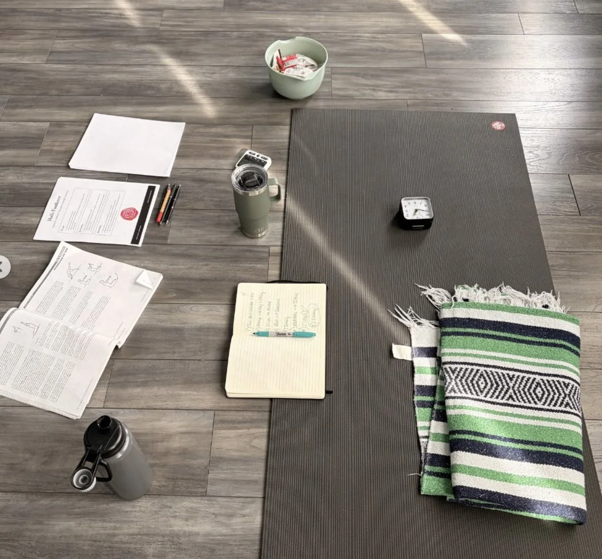 A brown yoga mat and folded Mexican blanket on a wooden floor with notebooks and coffee mugs.