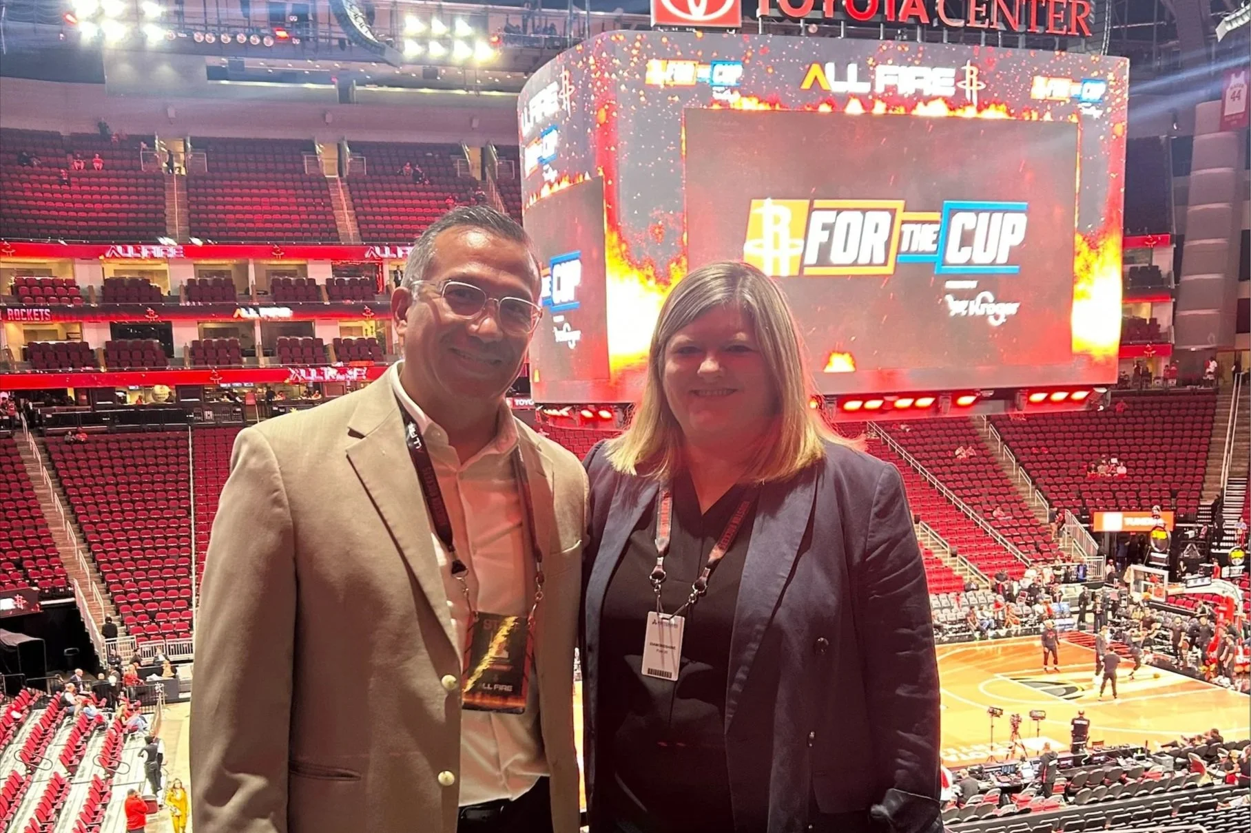 Two people standing in an arena with red seats and a basketball court, smiling at the camera. There is a large digital screen behind them displaying the event name 'ALL FIRE' with fire graphics and the phrase 'FOR THE CUP.' The arena is partially fil