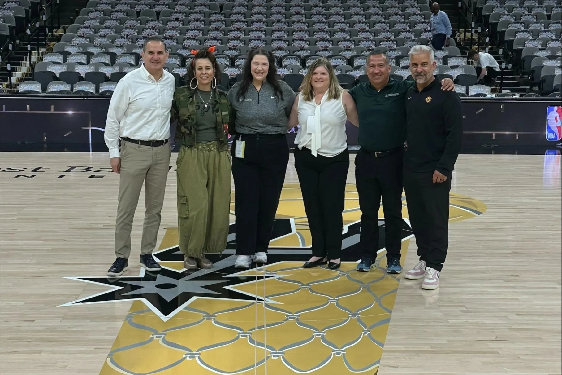 Group of six people standing on a basketball court, smiling at the camera. The court features a logo in the center and a black and gold design. Rows of empty chairs can be seen in the background, with a few people walking behind them.