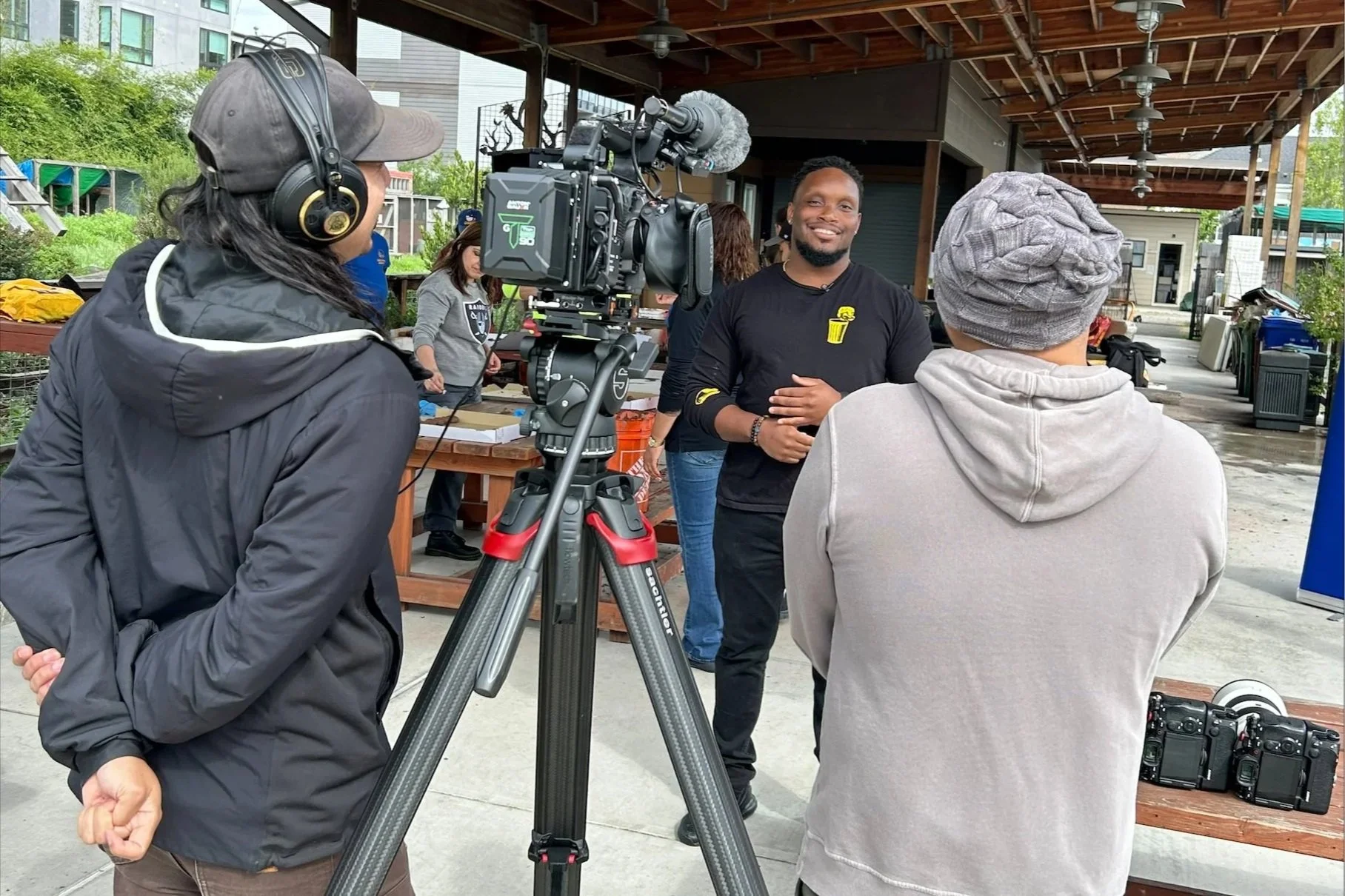 A person being filmed and interviewed outdoors, with a professional camera on a tripod aimed at him, while crew members listen and prepare equipment.